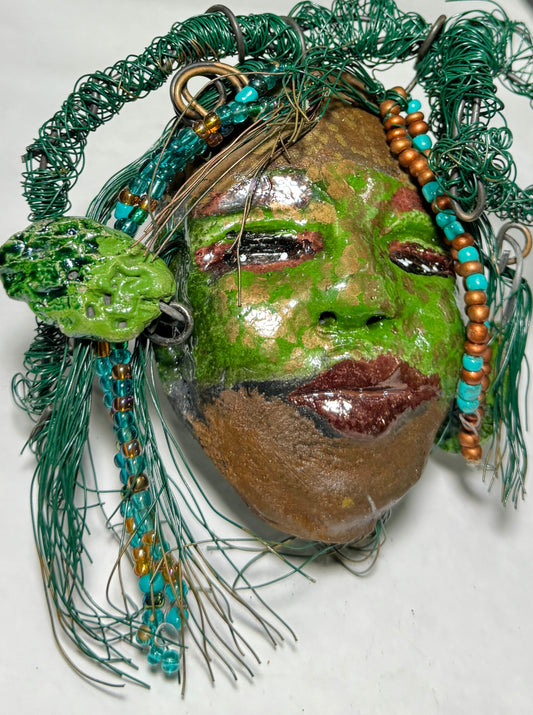 Taja is a Raku-fired mask measuring 4" x 5" x 2", featuring both 24 and 16 gauge wire to represent hair. This entrancing piece from the HerDew collection is the perfect gift for any holiday.