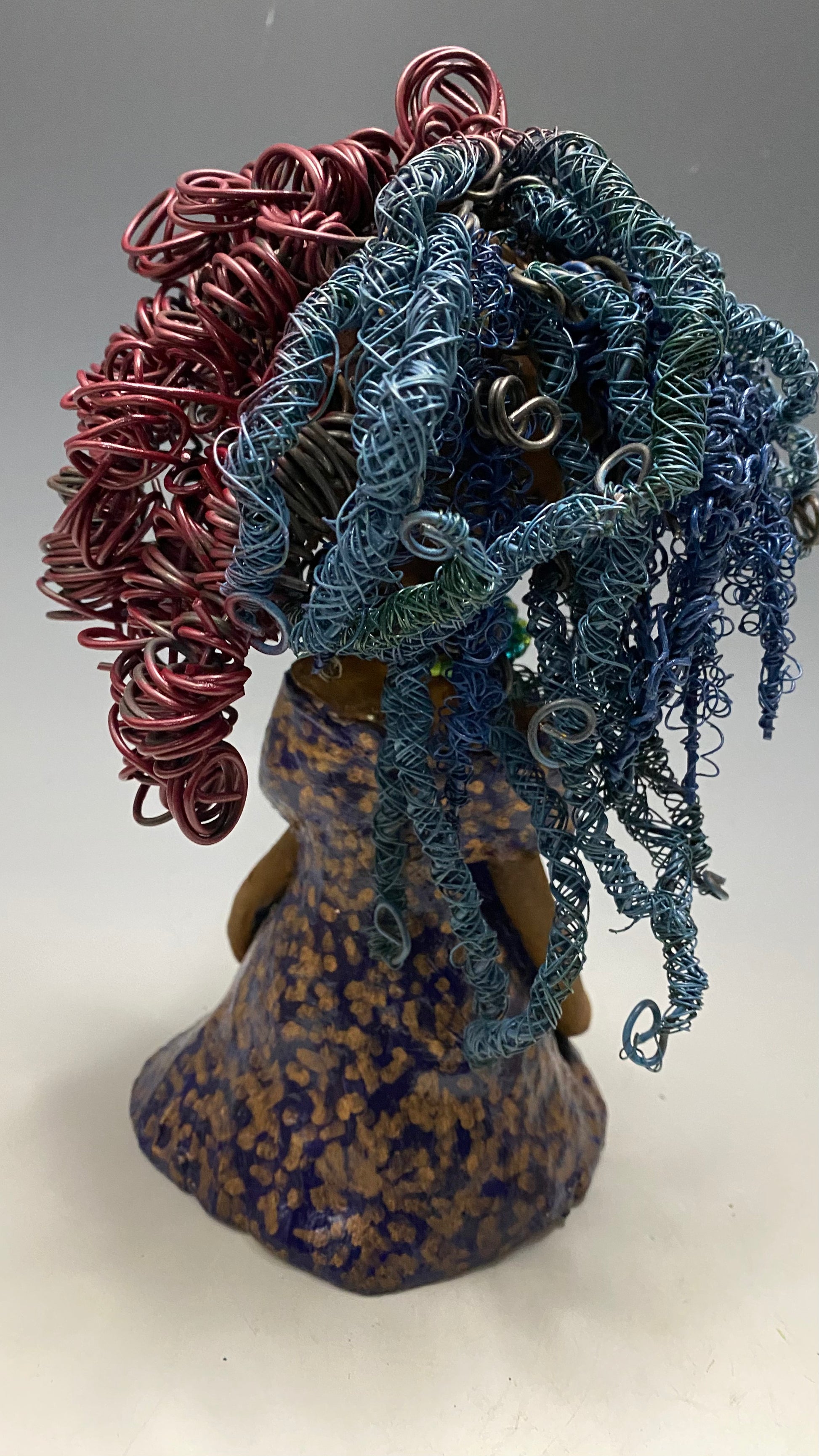Cristy is 9" x 6" x 5.5", weighs 2.5 lbs, and has a honey brown complexion with reddish brown lips. Her long hair is adorned with red and blue color and 80 feet of 24 and 16 gauge twisted wire. She wears an antique glazed dress featuring a spotted blue and gold pattern. Cristy is completed by her eyeglasses which sit upon her forehead, inspiring hope and sparking conversations. Visit us for questions