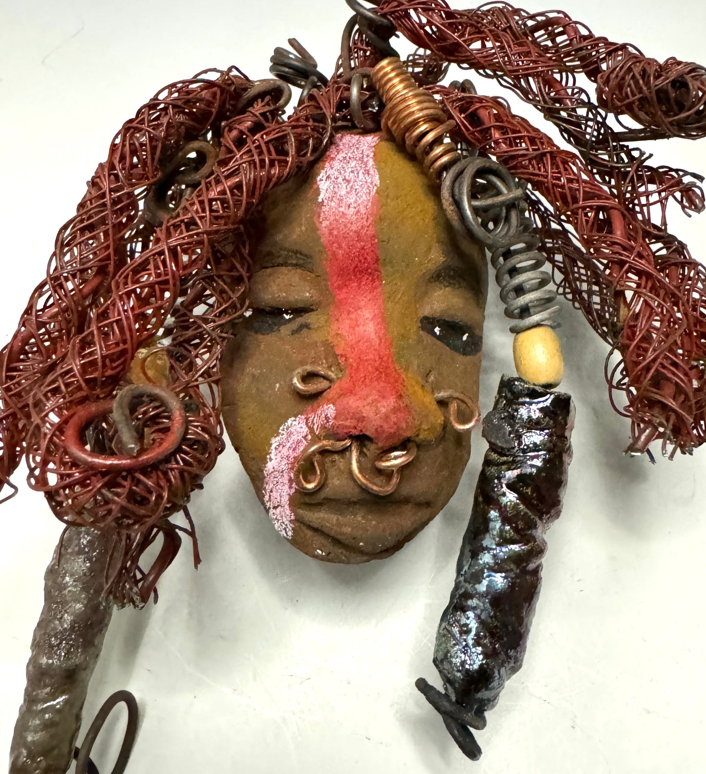 Billy is a 5" x 7" Raku-fired mask with intricate detail. Constructed with 16 and 24-gauge wire, spanning a total of 20 feet, he features a two tone, honey brown and red stripe complexion, matched with vibrant red coiled locs. Now available at a discount, Billy is an ideal choice for collectors or as a special holiday surprise.