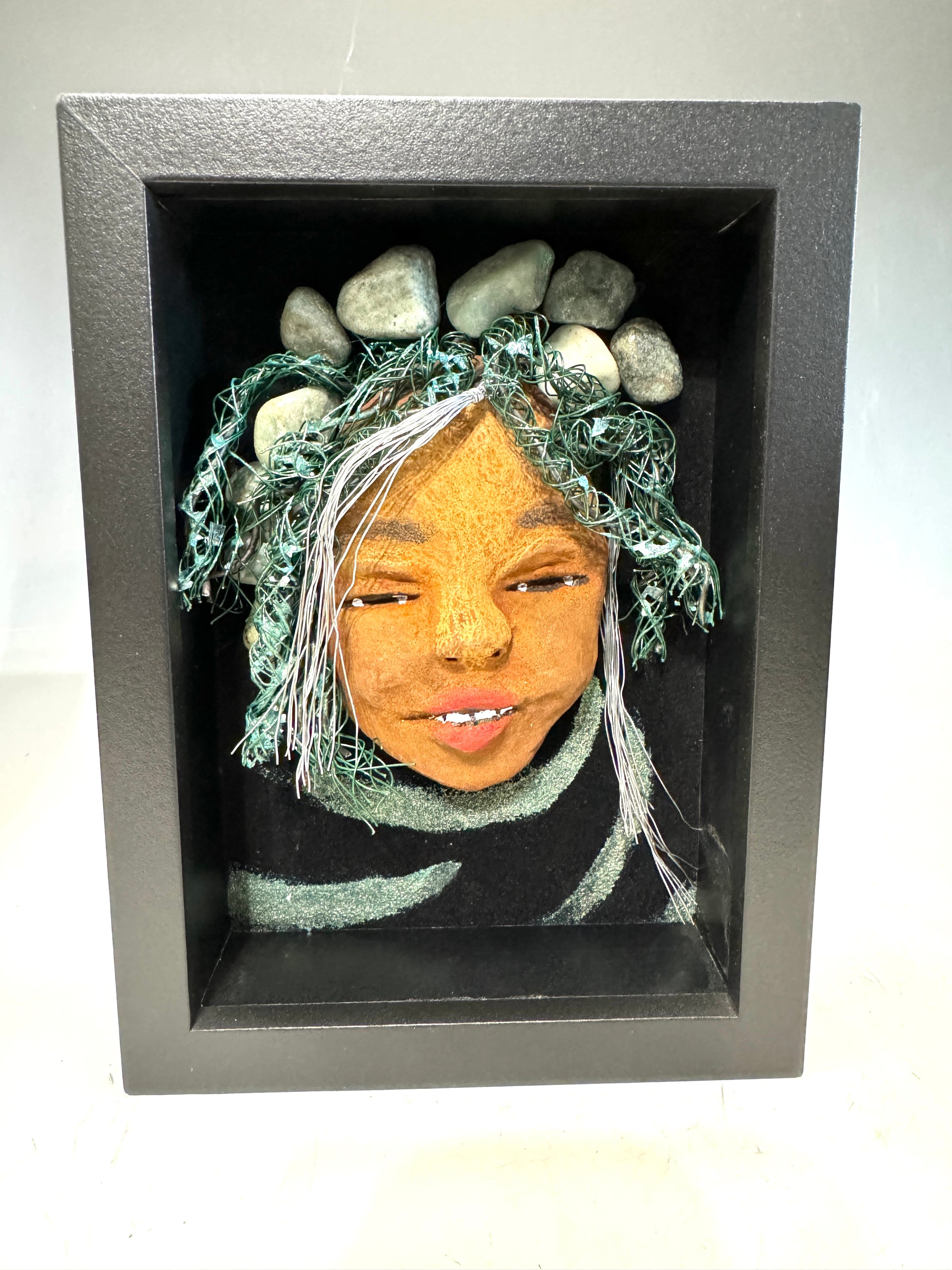 Introducing Brandie, a mixed media sculptured raku fired mask. Her features include a two-tone honey brown complexion, Ruby red lips, and hair intricately intertwined with 16 and 24 gauge wire. She is mounted on a hand-painted canvas, accented by an earthy landscape background, and enclosed in a 5“ x 7“ black shadowbox.