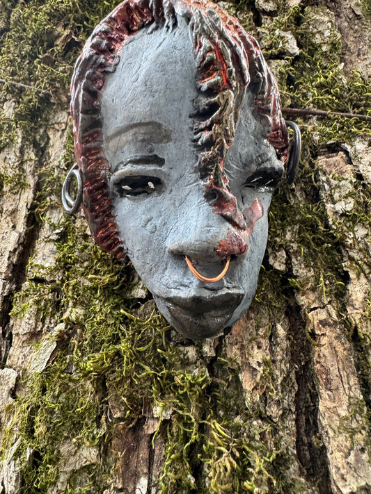 Tia weighs 3.2 oz and measures 4" by 3" in diameter. Her hair is copper-braided and her complexion is a light grayish blue hue with black lips. She is an ideal introductory piece from the HerDew mask collection.