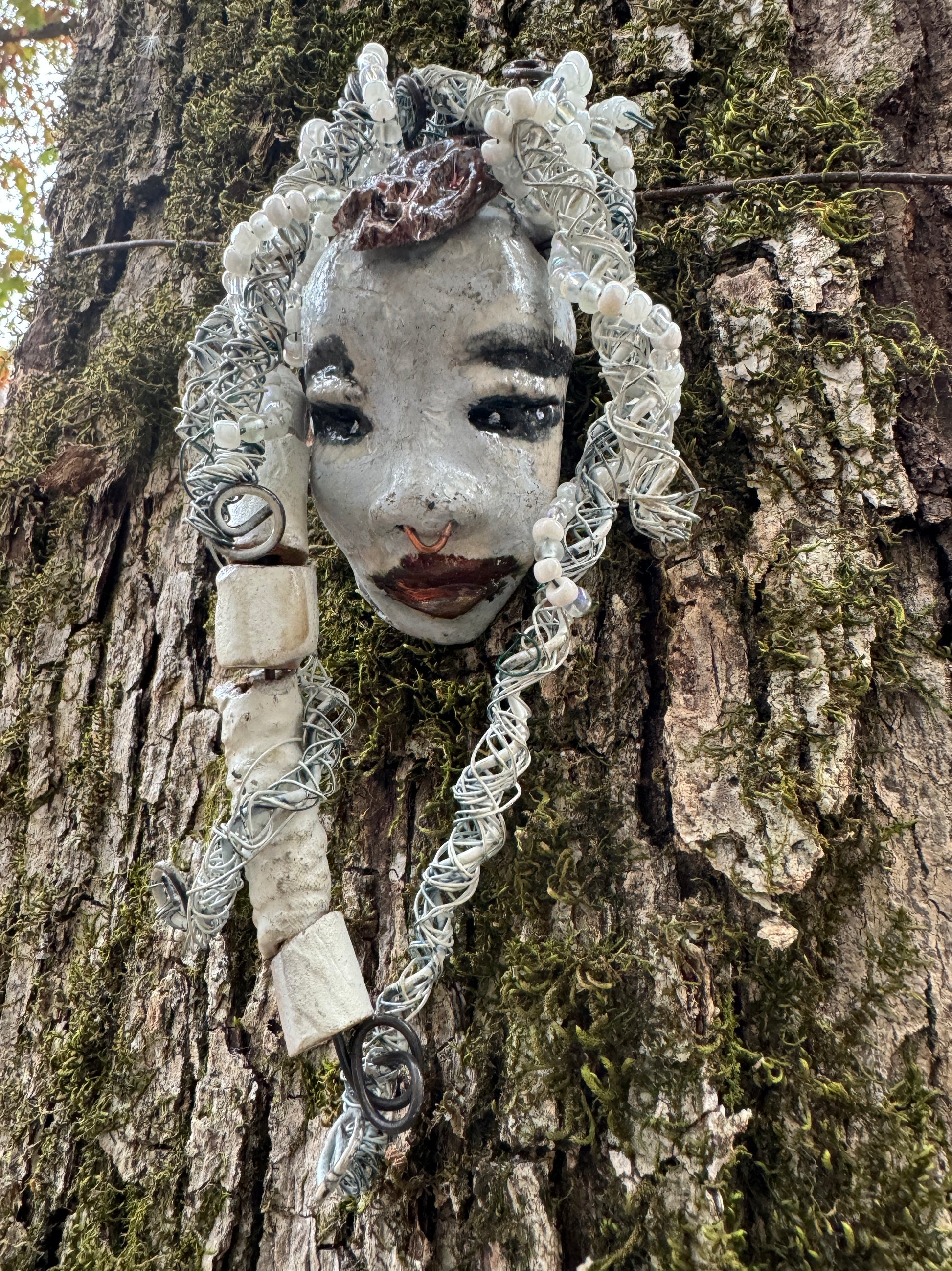 Faye is an absolutely stunning 4" x 4", 4.2 oz. Raku-fired mask with a white complexion, intricate beaded hair, and a one-of-a-kind portrait setting of a majestic tree. She's sure to make a statement indoors or outdoors - an artful gift for nature admirers and décor fans alike!
