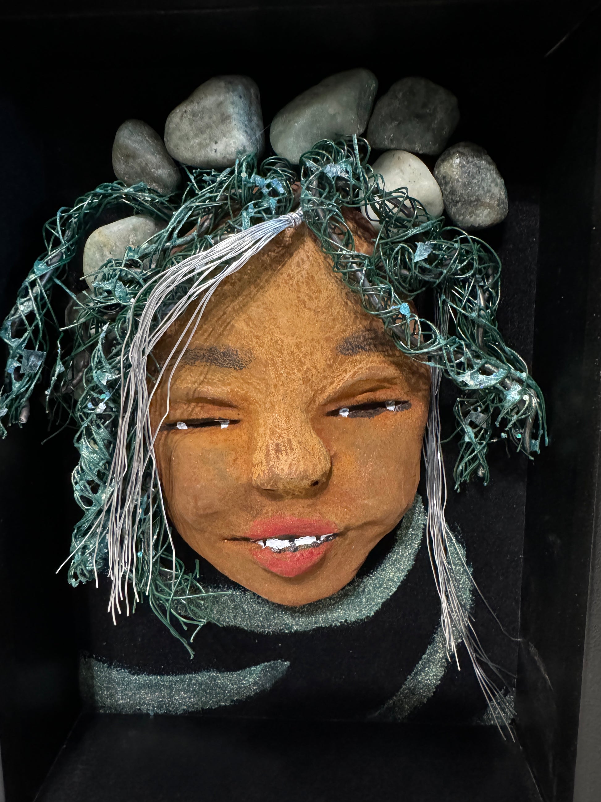 Introducing Brandie, a mixed media sculptured raku fired mask. Her features include a two-tone honey brown complexion, Ruby red lips, and hair intricately intertwined with 16 and 24 gauge wire. She is mounted on a hand-painted canvas, accented by an earthy landscape background, and enclosed in a 5“ x 7“ black shadowbox.
