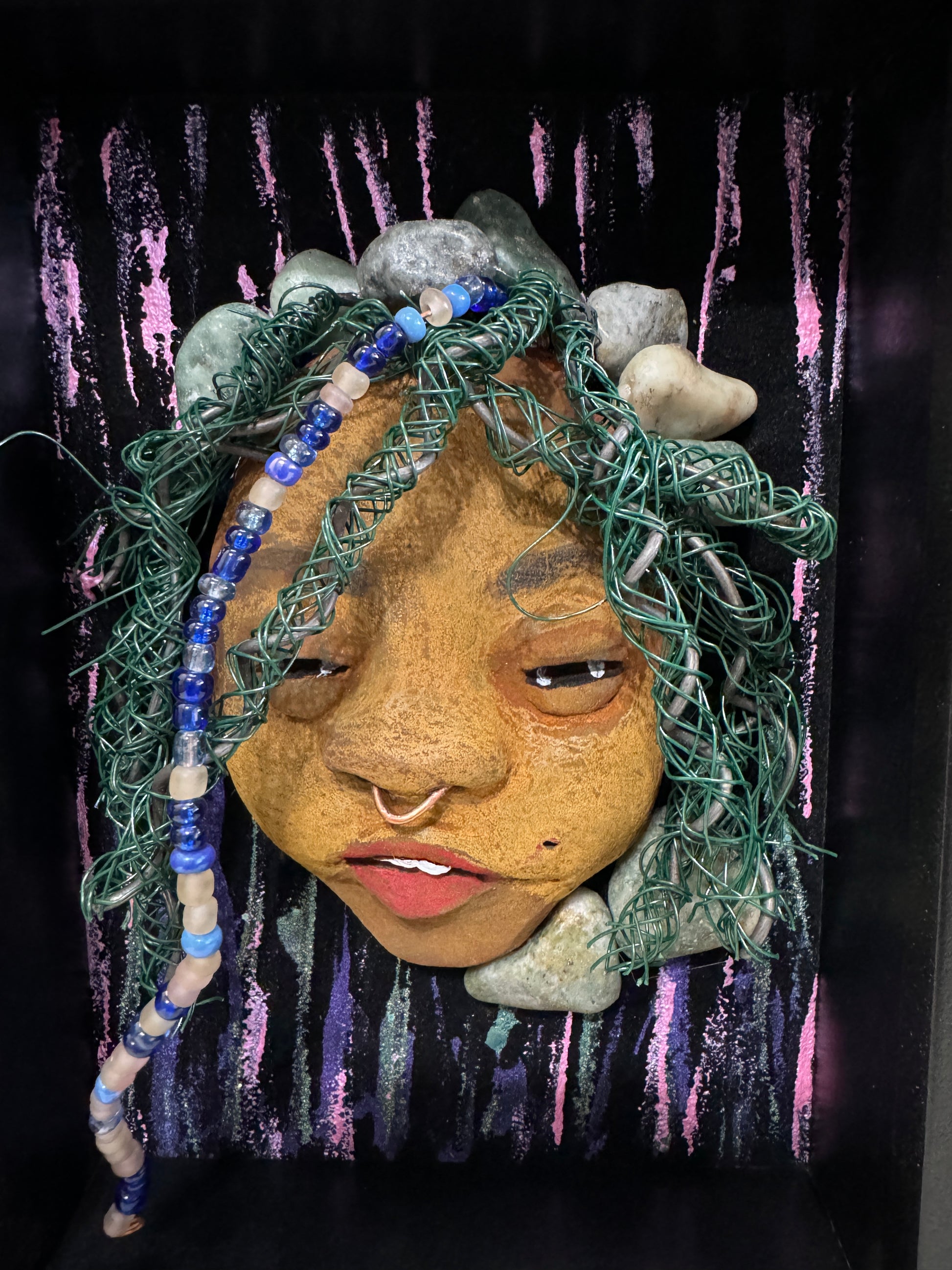Introducing Beverly: A striking mixed media mask created using the raku firing method. The meticulously designed features showcase a beautiful honey brown complexion with subtle Ruby red lips and intricately woven hair made with 16 and 24 gauge wire. This exquisite piece is mounted on a hand-painted canvas, featuring a stunningly landscaped background, and elegantly presented in a glass covered 5" x 7" black shadowbox.