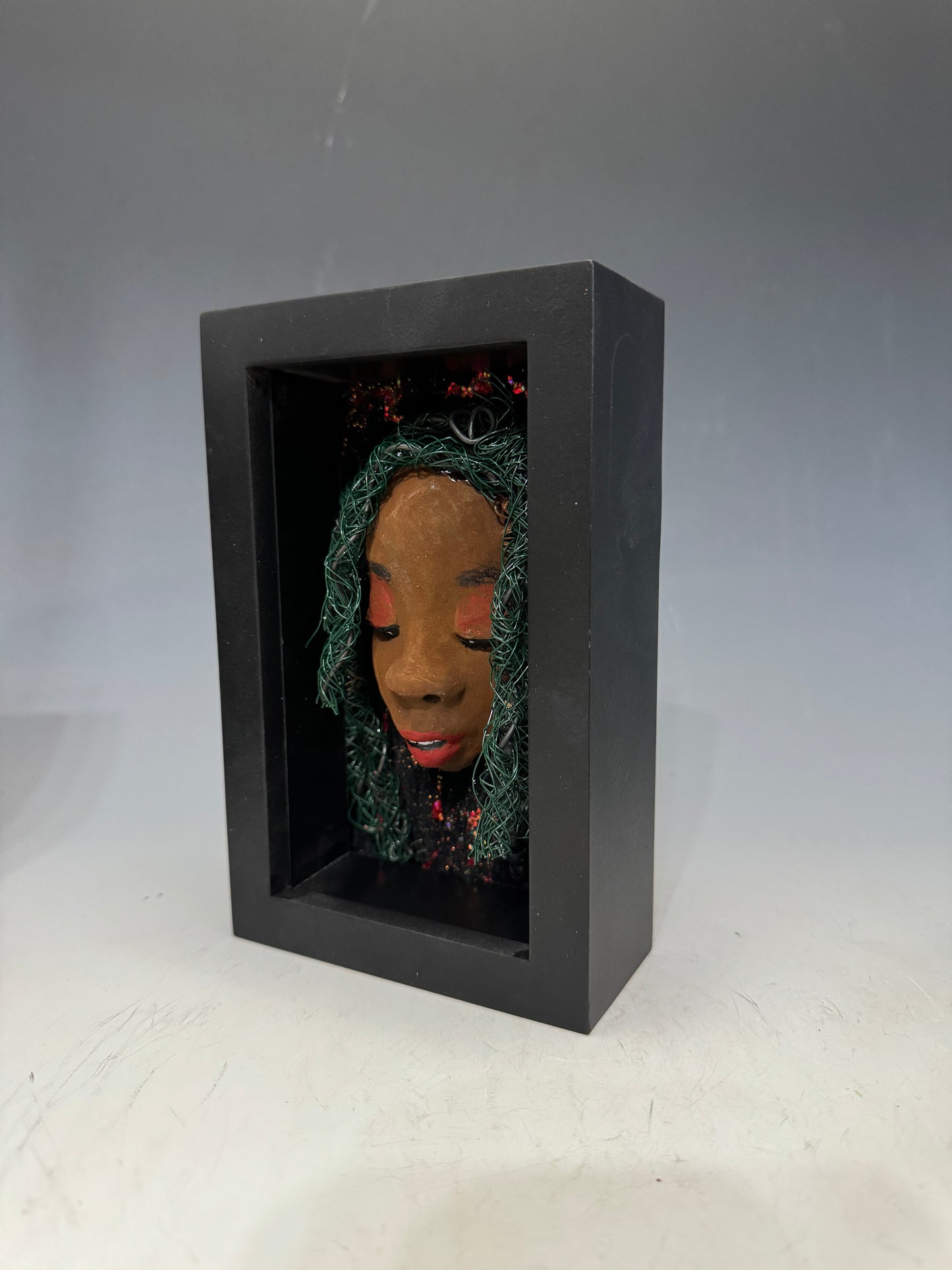 Marjorie is a mixed media mask crafted with the raku firing technique, displaying carefully detailed characteristics such as honey brown skin, Ruby red lips, and intricately woven hair made from 16 and 24-gauge wire. This stunning piece is mounted on a hand-painted canvas, surrounded by a beautifully landscaped background, and elegantly displayed in a glass-covered 4" x 6" black shadowbox.