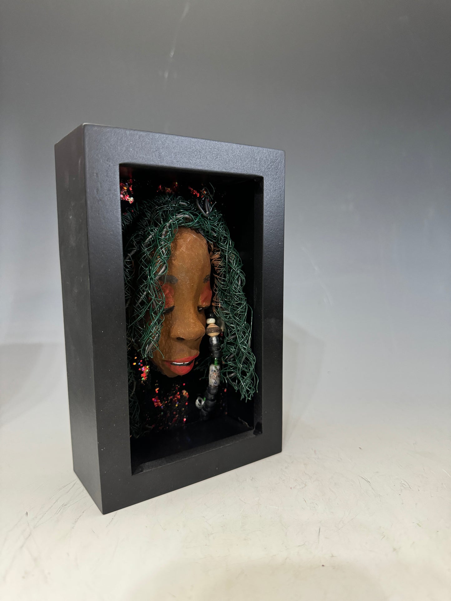 Marjorie is a mixed media mask crafted with the raku firing technique, displaying carefully detailed characteristics such as honey brown skin, Ruby red lips, and intricately woven hair made from 16 and 24-gauge wire. This stunning piece is mounted on a hand-painted canvas, surrounded by a beautifully landscaped background, and elegantly displayed in a glass-covered 4" x 6" black shadowbox.