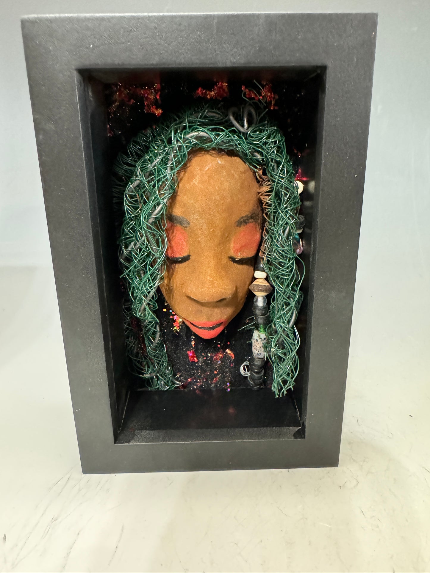 Marjorie is a mixed media mask crafted with the raku firing technique, displaying carefully detailed characteristics such as honey brown skin, Ruby red lips, and intricately woven hair made from 16 and 24-gauge wire. This stunning piece is mounted on a hand-painted canvas, surrounded by a beautifully landscaped background, and elegantly displayed in a glass-covered 4" x 6" black shadowbox.