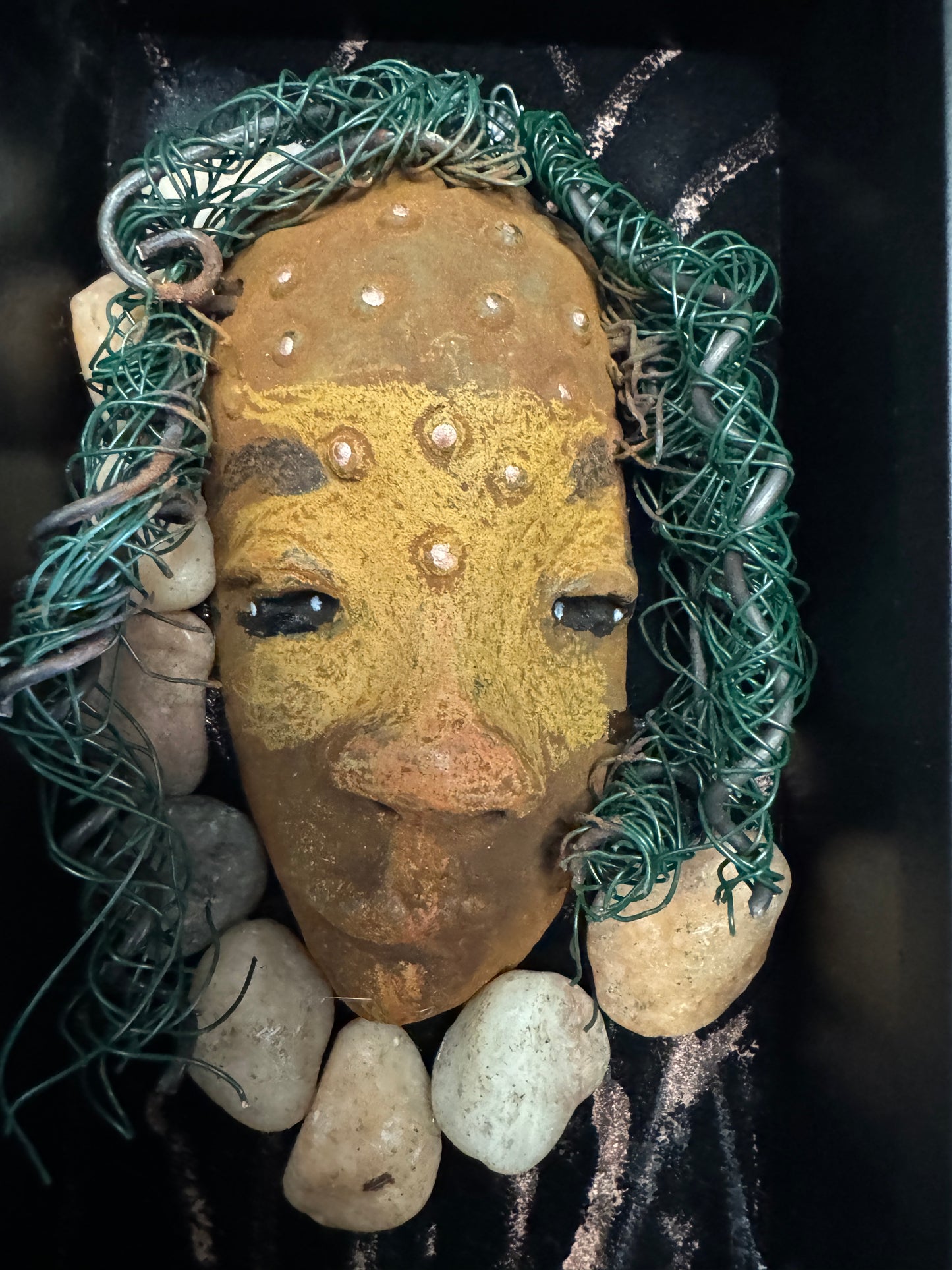 Presenting Craig: A captivating mixed media mask made with a raku firing technique. Meticulously crafted features include a unique two-tone honey brown complexion, subtle rust brown red lips, and hair intricately woven with 16 and 24 gauge wire. This piece is mounted on a hand-painted canvas, featuring a beautifully landscaped background, and elegantly displayed in a glass covered 4" x 6" black shadowbox.