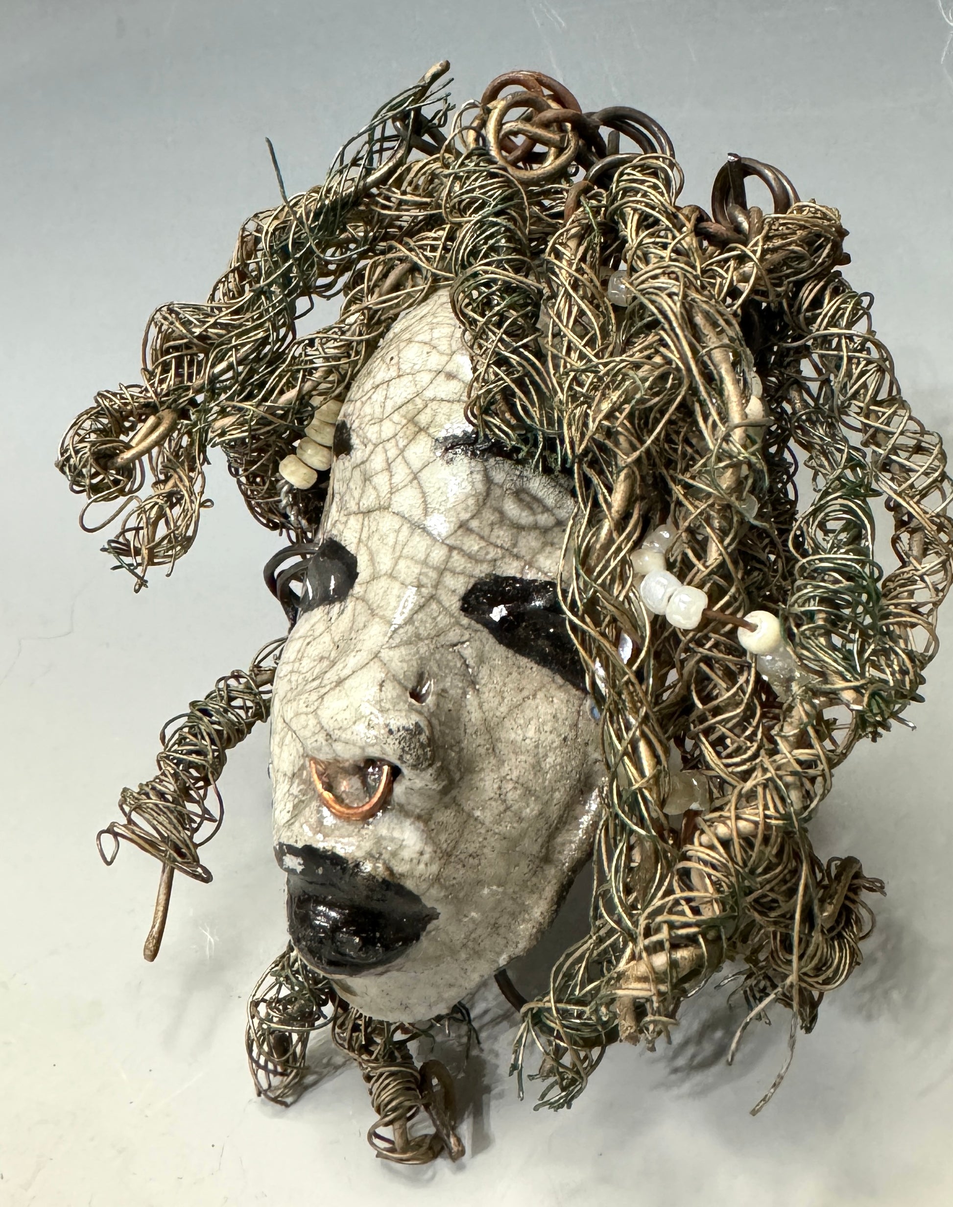 ami is a 4x 6” raku-fired mask featuring a pristine off-white color palette. Her intricately detailed hair is composed of nearly 15 feet of 16 gauge wire, delicately adorned with dainty beads. Set in a backdrop of nature, her portrait makes a wonderful addition to any outdoor art collector, or for decorating the home.
