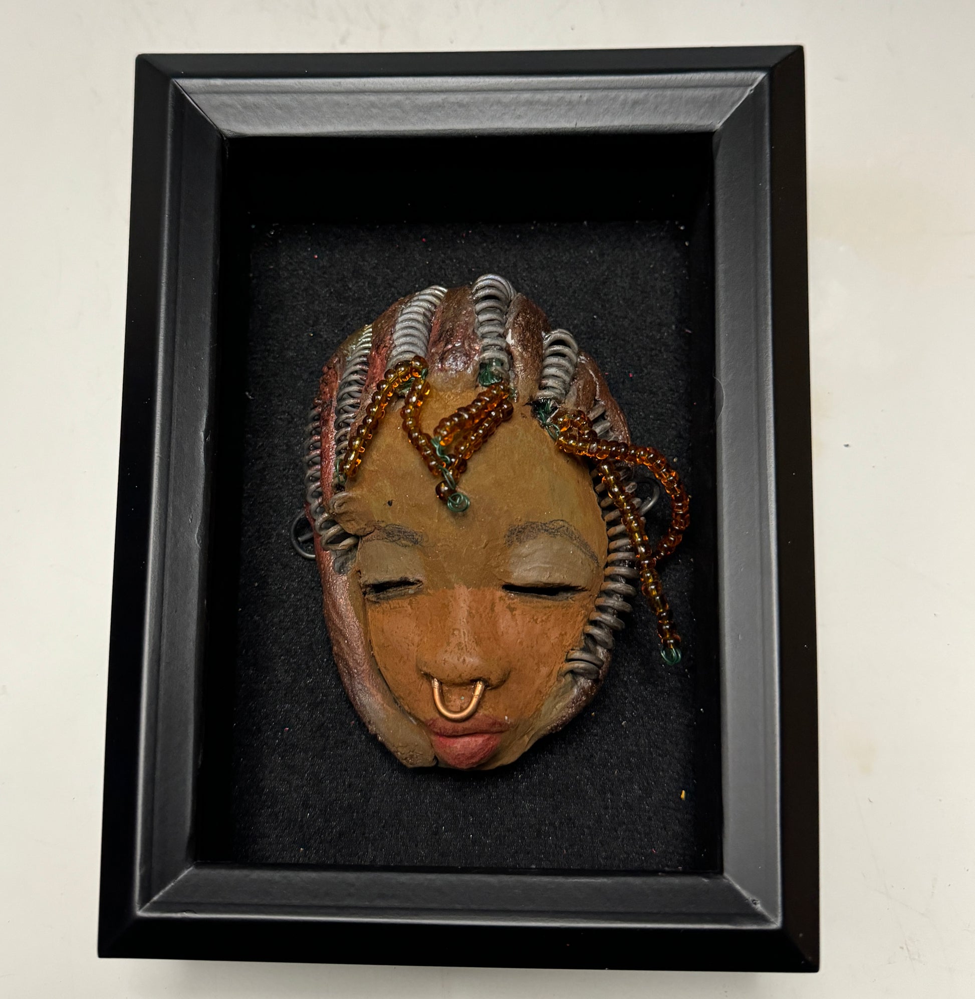Kenya weighs 5 oz and is 4" by 3" in diameter. Her hair is coppered braided, her complexion is a light gray-blue, and her lips are black. She is a perfect item to start with from the HerDew mask collection. Shadowbox not included.