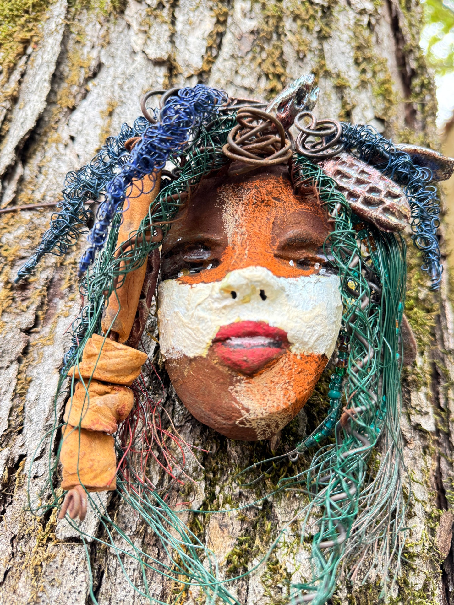“Malik Trueborn” — This striking 5” x 4” mask weighs 8 ozs. It is a testament to resilience and identity, crafted with both 16 and 24 gauge wire, creating a powerful yet delicate framework. The intricate claywork with mixed elements form abstract contours and expressive features, capturing a sense of strength and mystery in its compact form. Accented by layers of raku fire glazes, the mask shimmers with unpredictable swirls of earthy coppers, iridescent blues, and deep, smoky blacks, giving it a