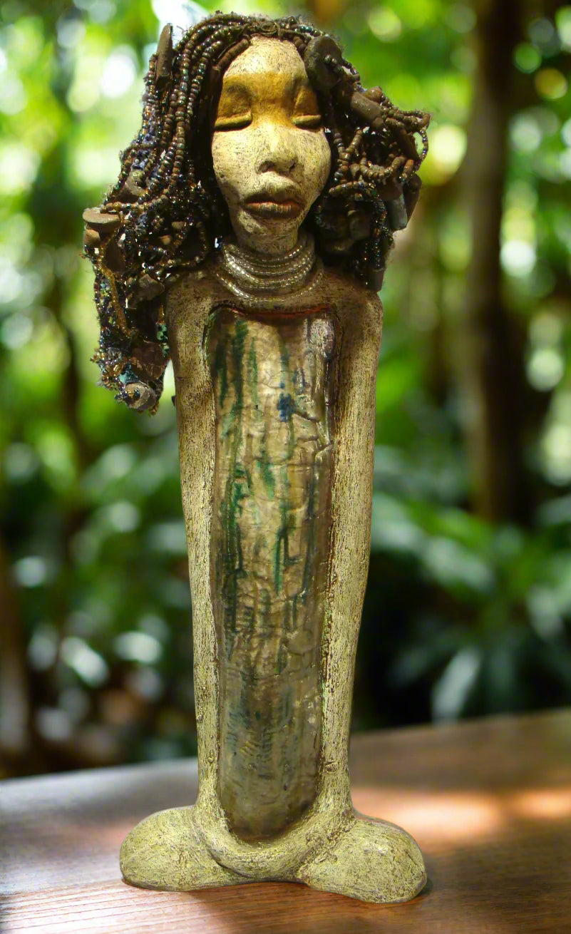 Meet Ariel, a meticulously crafted beauty. Standing at 21" x 9" x 4.5" and weighing 7lbs, she boasts a stunning beige and honey brown complexion. Her head is adorned with a variety of seed, pony, and raku beaded hair, adding to her allure. Adorned in a metallic alligator green dress with hints of shimmering copper, Ariel commands attention with her exotic gaze.