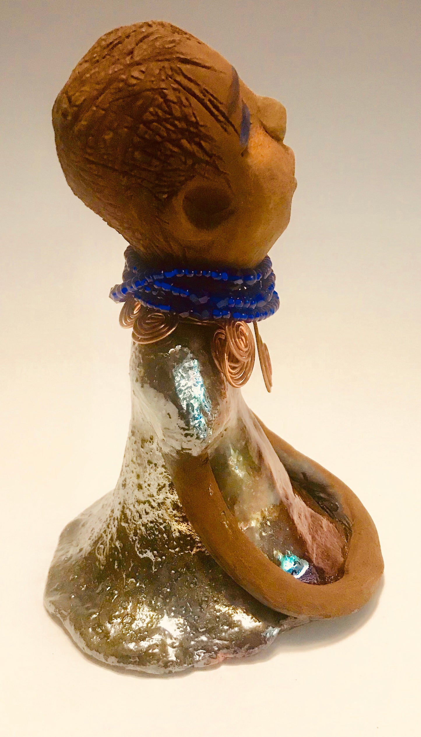 Sade stands 7.5" x 5" x 5" and weighs 1.04 lbs. She has a lovely honey brown complexion with reddish brown lips. She has a short braided hairstyle. Sade has a colorful metallic antique copper glazed dress. She wears spiral copper wire necklaces on top of an aqua blue beaded collar. Sade long loving arms rest at her side. With accent blue eye shadow and eyes wide opened, Sade has hopes of finding a new home.