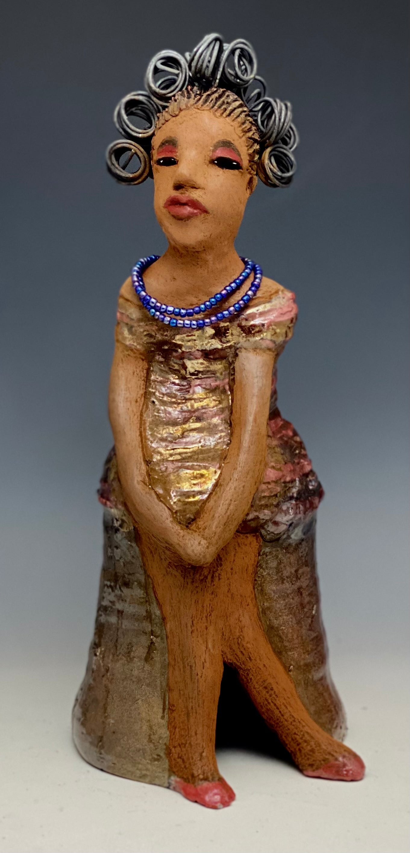 You know someone like Kim. One who reaps elegance and sophistication! Kim stands 13" x 5" x 7" and weighs 2.07 lbs. She has a lovely light beige complexion with over 4 feet of 16 gauge spiral coiled wire hair. Kim's metallic copper red and gold dress is accented by a multicolored aqua blue beaded necklace. Kim's maroon eye shadow gives her a distinguished mystical look. One must wonder what is Kim thinking? Kim sits with her legs crossed as she awaits a special place to grace your home.