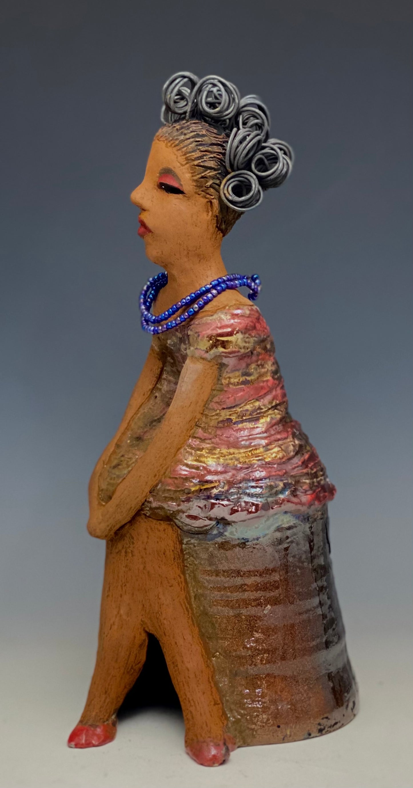 You know someone like Kim. One who reaps elegance and sophistication! Kim stands 13" x 5" x 7" and weighs 2.07 lbs. She has a lovely light beige complexion with over 4 feet of 16 gauge spiral coiled wire hair. Kim's metallic copper red and gold dress is accented by a multicolored aqua blue beaded necklace. Kim's maroon eye shadow gives her a distinguished mystical look. One must wonder what is Kim thinking? Kim sits with her legs crossed as she awaits a special place to grace your home.