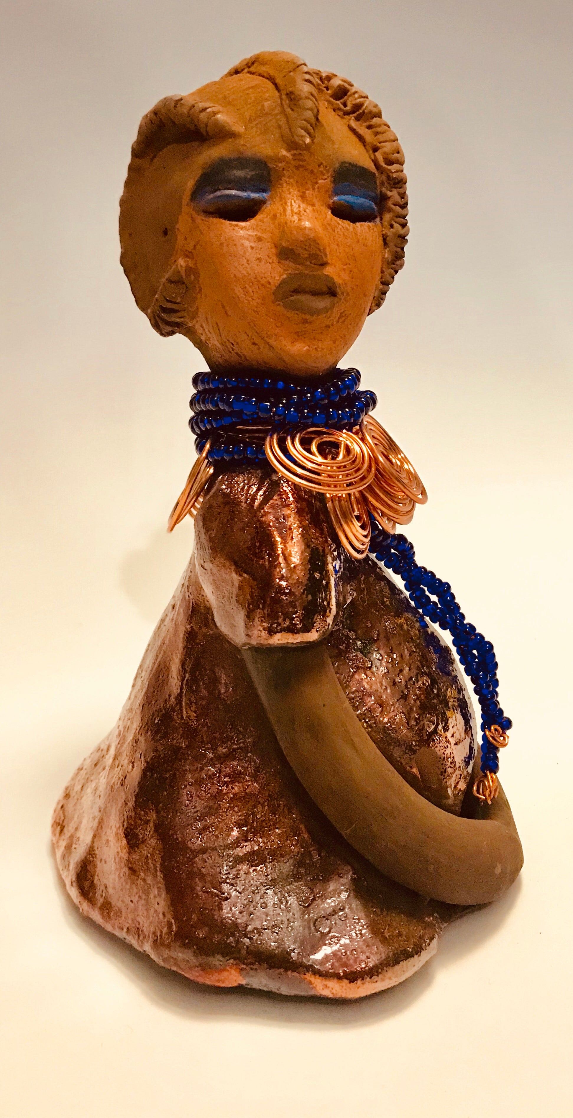Ife stands 7.5" x 4" x 5" and weighs 1.02  lbs. She has a lovely honey brown complexion with reddish brown lips. She has a short braided hairstyle.      Ife has a colorful metallic antique copper glazed dress.     She wears spiral copper wire necklaces on top of an aqua blue beaded collar.     Ife long loving arms rest at her side.     With accent blue eye shadow and eyes wide opened, Ife has hopes of finding a new home.