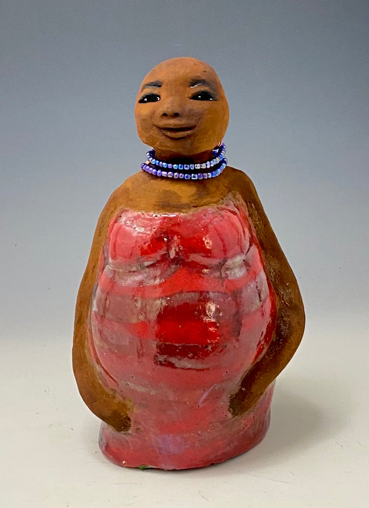 Meet Cora! Cora stands 9" x 7" x 4.5" and weighs 2lbs. She has a lovely honey brown complexion with lovely reddish brown lips. Cora wears a clay bald hairstyle. Cora has a striped heart red and gold glazed dress. She wears a blue beaded necklace. With eyes wide opened, Janae' has hopes of finding a new home.