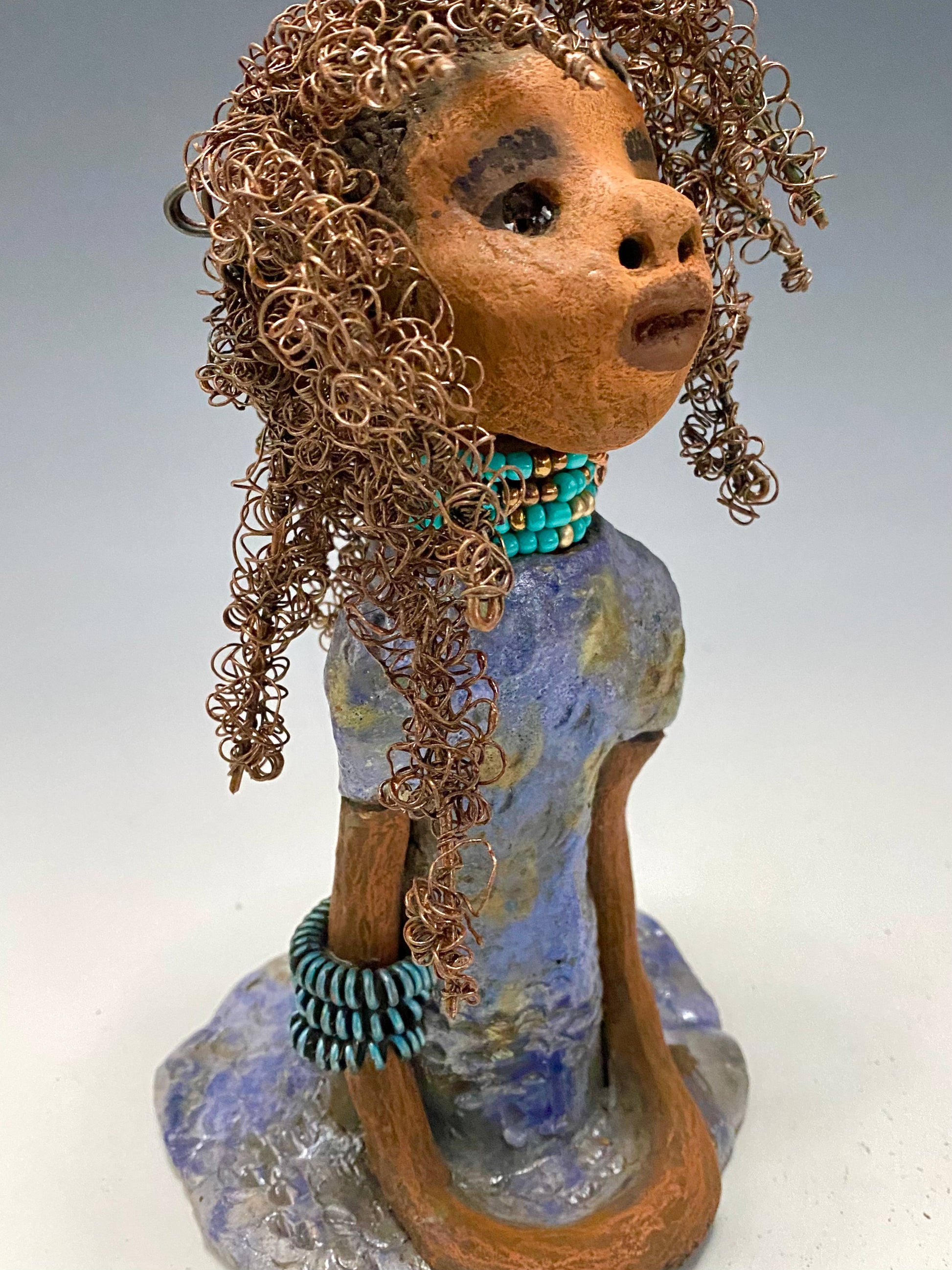 Meet Janae'! She has grown a head full of Hair! Janae' stands 8" x 5" x 4" and weighs 1.03 lbs. She has a lovely honey brown complexion with lovely reddish brown lips. Janae' wears a clay braided hairstyle. Janae' has a colorful metallic blue and gold glazed dress. She wears a blue and antique copper  beaded necklace. With eyes wide opened, Janae' has hopes of finding a new home.