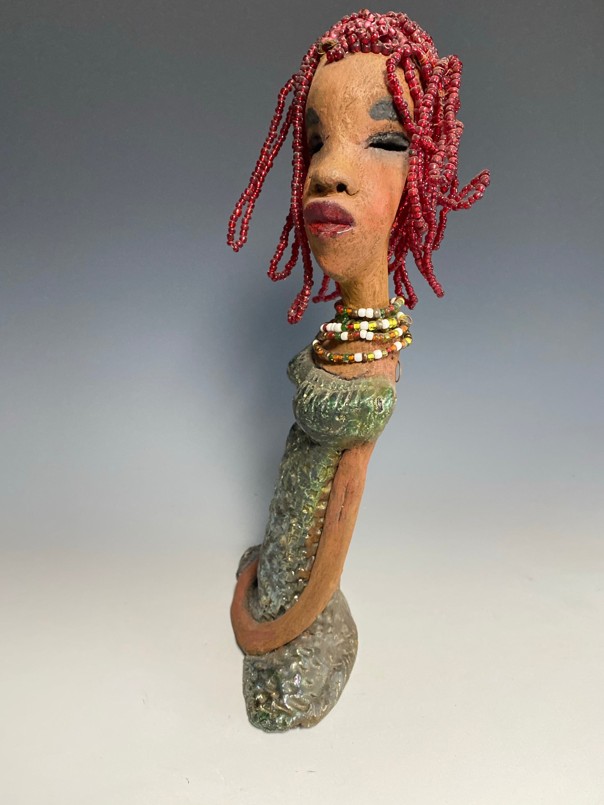 Meet July! July stands 14" x 5" x 3" and weighs 2.05 lbs. July has a head full of red beaded hair. She has a honey brown complexion. July dress has a metallic textured copper green.  She wears a multi colored beaded necklace. With her head slightly turned her long loving arms gently rest at her side.