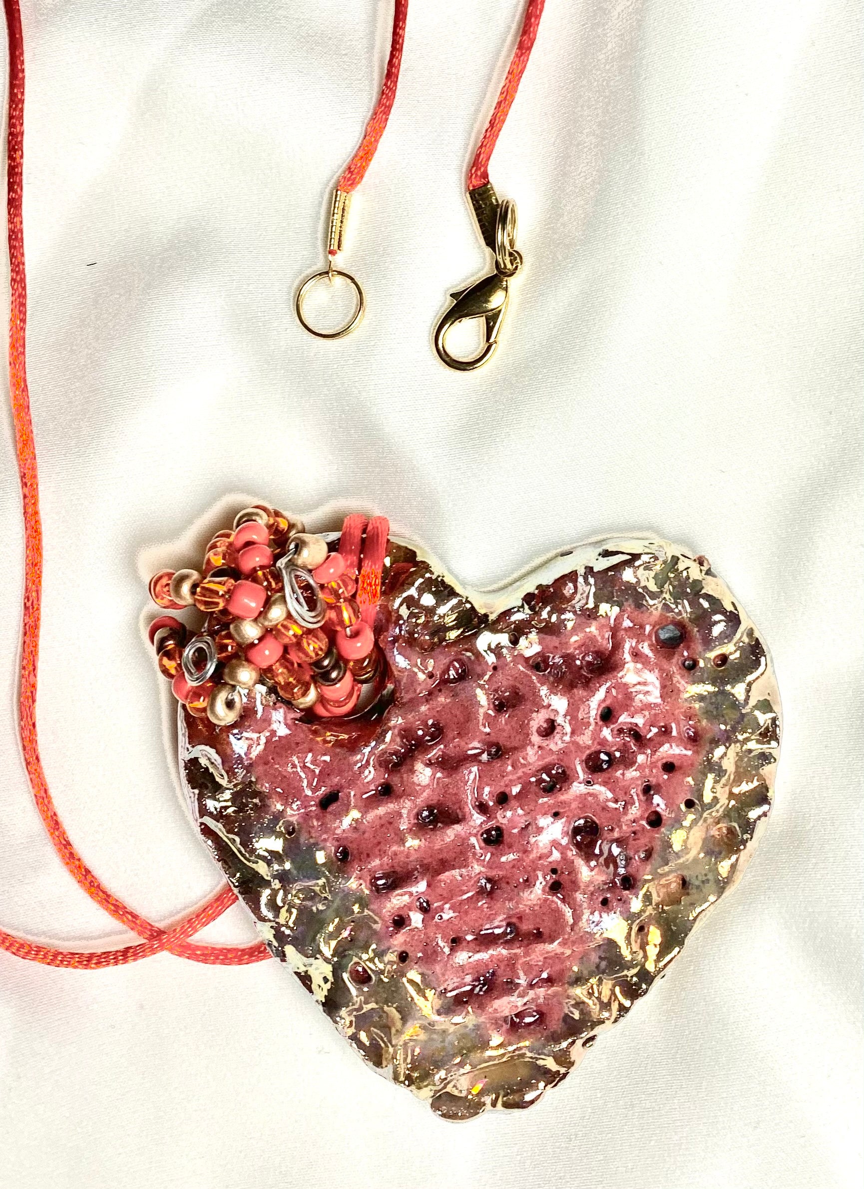Have A Heart ! Each heart pendant is handmade with love! It is 3"x 3" and weighs approx. 3ozs. This pendant has a red violet and gold metallic raku glazes that renders a unique translucent  patina. The heart has a textured pattern . Both sides are  are different and equally beautiful! It holds a spiral of red and gold mini beads on a spiral copper wire. This pendant has a nice 12" red rattail cord!