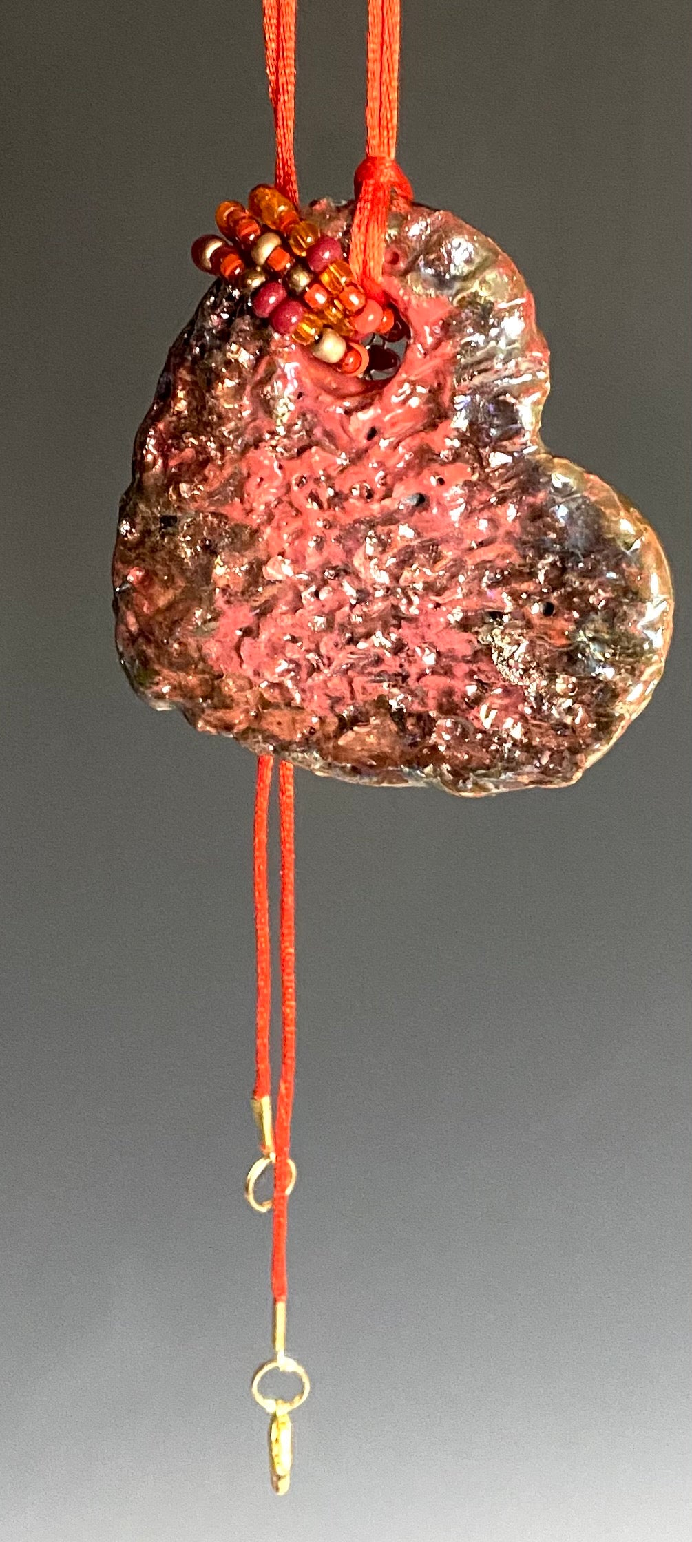 Have A Heart! Each heart pendant is handmade with love! It is 3"x 3" and weighs approx. 3ozs. This pendant has a red violet and gold metallic raku glazes that renders a unique translucent patina. The heart has a textured pattern . Both sides are are different and equally beautiful! It holds a spiral of red mini beads on a spiral copper wire. This pendant has a nice 12" red rattail cord!