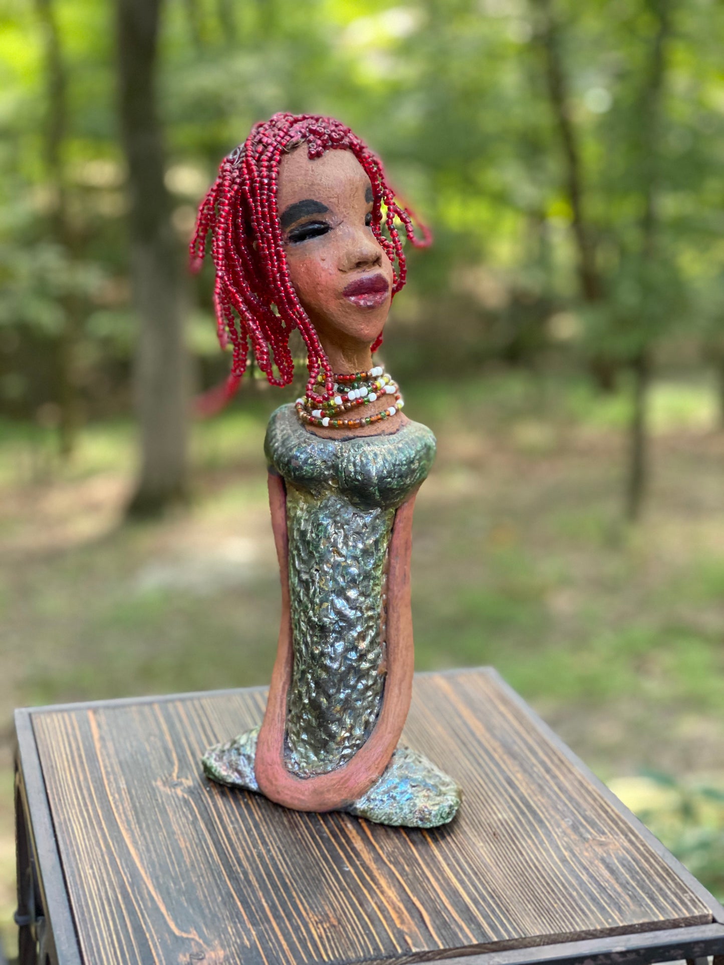 Meet July! July stands 14" x 5" x 3" and weighs 2.05 lbs. July has a head full of red beaded hair. She has a honey brown complexion. July dress has a metallic textured copper green. She wears a multi colored beaded necklace. With her head slightly turned her long loving arms gently rest at her side.