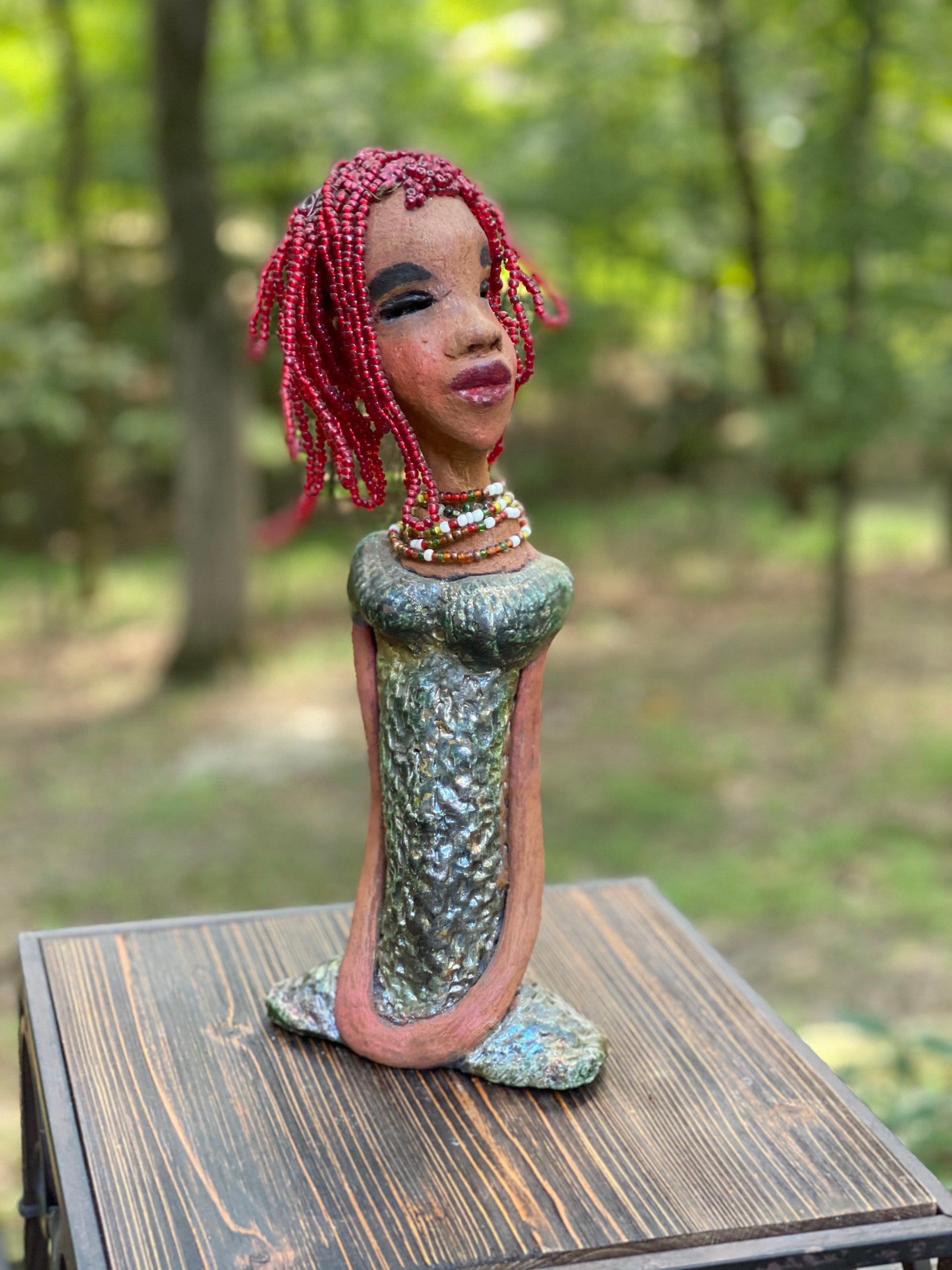 Meet July! July stands 14" x 5" x 3" and weighs 2.05 lbs. July has a head full of red beaded hair. She has a honey brown complexion. July dress has a metallic textured copper green.  She wears a multi colored beaded necklace. With her head slightly turned her long loving arms gently rest at her side.