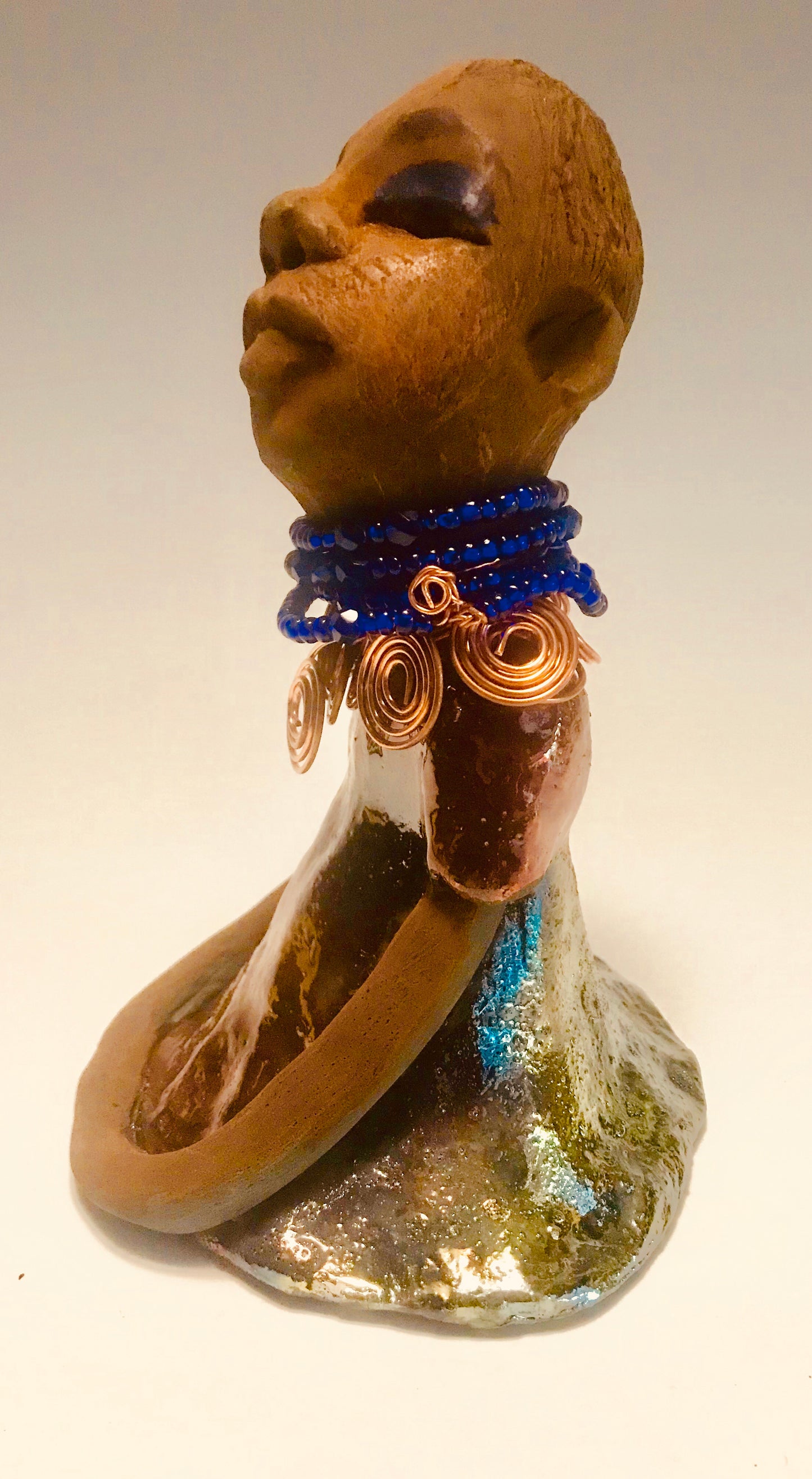 Sade stands 7.5" x 5" x 5" and weighs 1.04 lbs. She has a lovely honey brown complexion with reddish brown lips. She has a short braided hairstyle. Sade has a colorful metallic antique copper glazed dress. She wears spiral copper wire necklaces on top of an aqua blue beaded collar. Sade long loving arms rest at her side. With accent blue eye shadow and eyes wide opened, Sade has hopes of finding a new home.