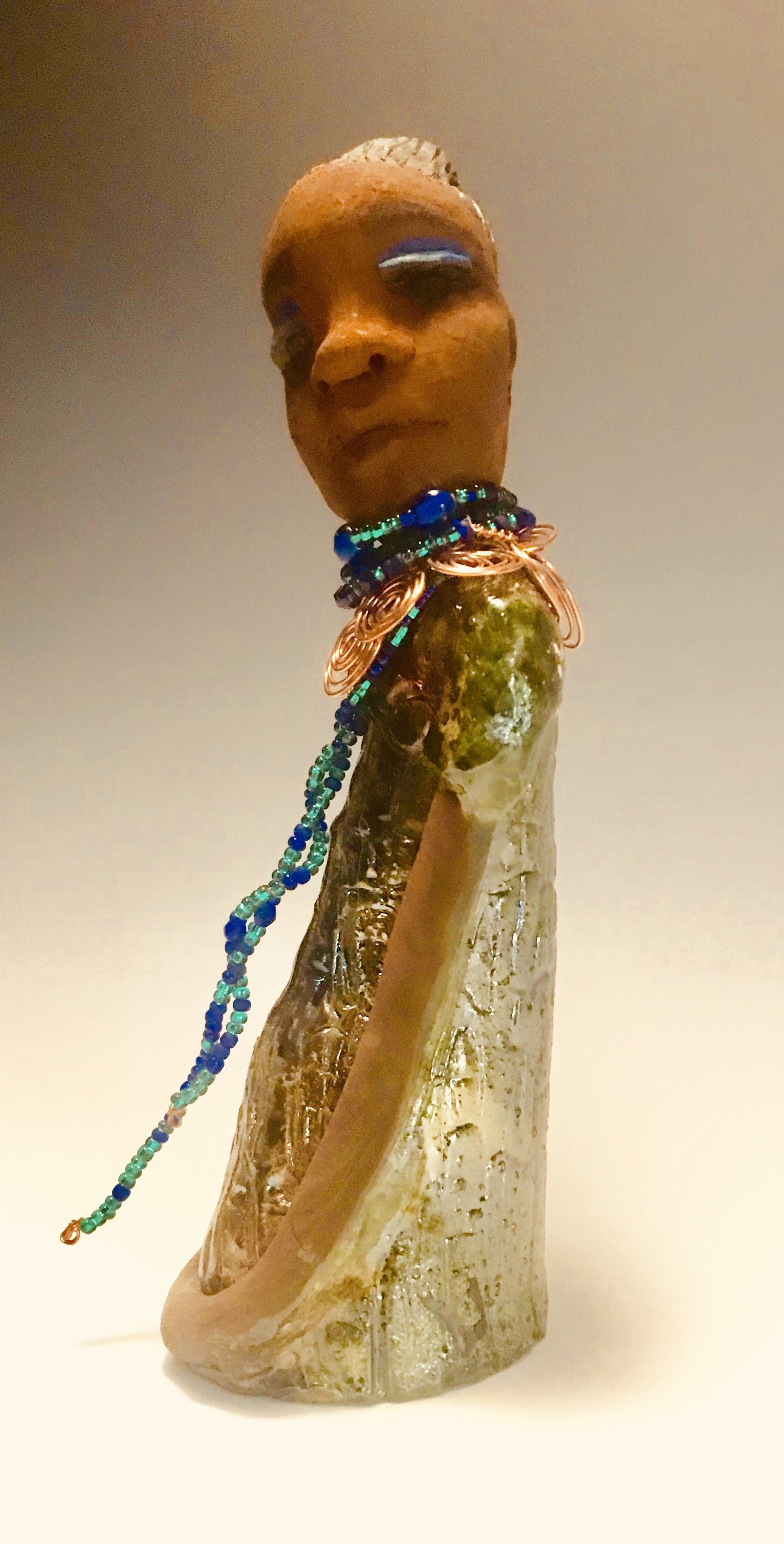 Emma stands 12.5" x3. 5" x 4" and weighs 2.04 lbs. She has a lovely honey brown complexion with reddish brown lips. She has a braided hairstyle. Emma has a colorful metallic green antique copper glazed dress. She wears a spiral copper wire necklace on top of a multicolored blue beaded collar. With eyes wide opened, Emma has hopes of finding a new home.