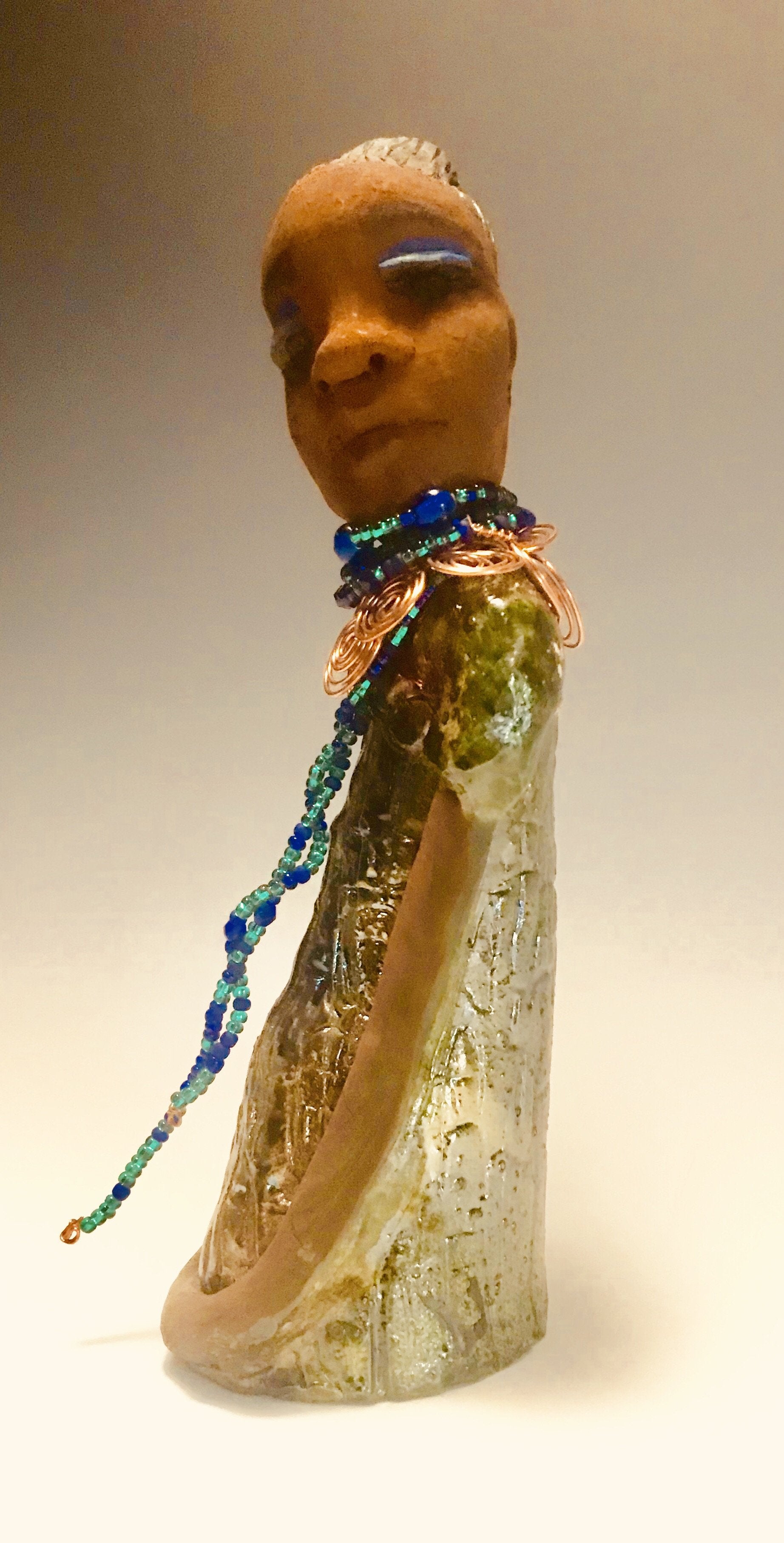 Emma stands 12.5" x3. 5" x 4" and weighs 2.04 lbs. She has a lovely honey brown complexion with  reddish brown lips. She has a braided hairstyle.  Emma has a colorful metallic green antique copper glazed dress. She wears a spiral copper wire necklace on top of a multicolored blue beaded collar. With eyes wide opened,  Emma has hopes of finding a new home.