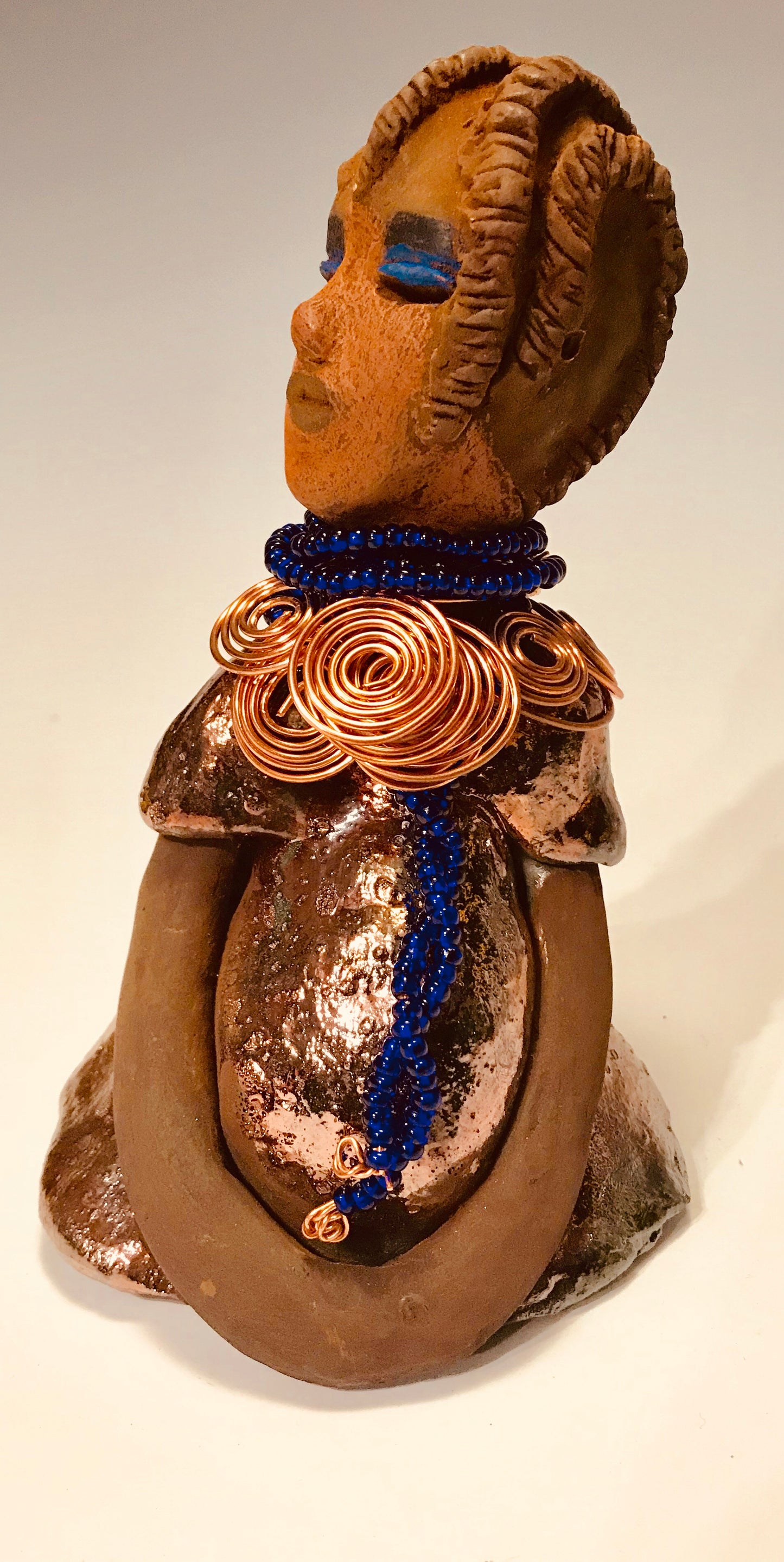Ife stands 7.5" x 4" x 5" and weighs 1.02 lbs. She has a lovely honey brown complexion with reddish brown lips. She has a short braided hairstyle. Ife has a colorful metallic antique copper glazed dress. She wears spiral copper wire necklaces on top of an aqua blue beaded collar. Ife long loving arms rest at her side. With accent blue eye shadow and eyes wide opened, Ife has hopes of finding a new home.