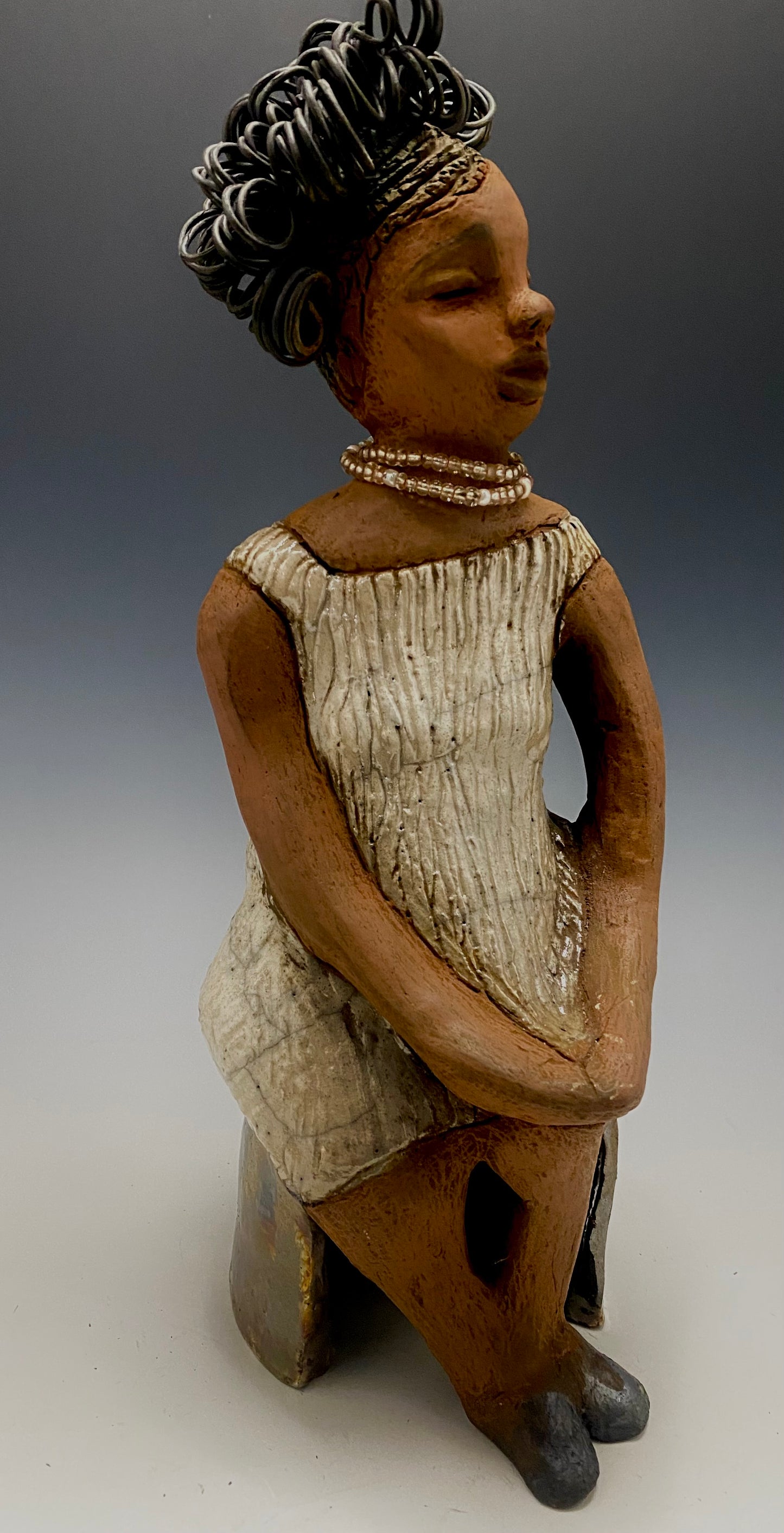 Meet Gwen! You know someone like Gwen. One who reaps elegance and sophistication! Gwen stands 13 " x 6" x 6" and weighs 3.07 lbs. She has a lovely brown complexion with over 5 feet of 16 gauge spiral coiled wire hair. Gwen's metallic off white dress is accented by a multicolored white and amber beaded necklace. Gwen’s straight face gives her a distinguished no nonsense look. Gwen sits with her hands rested as she awaits a special place to grace your home.