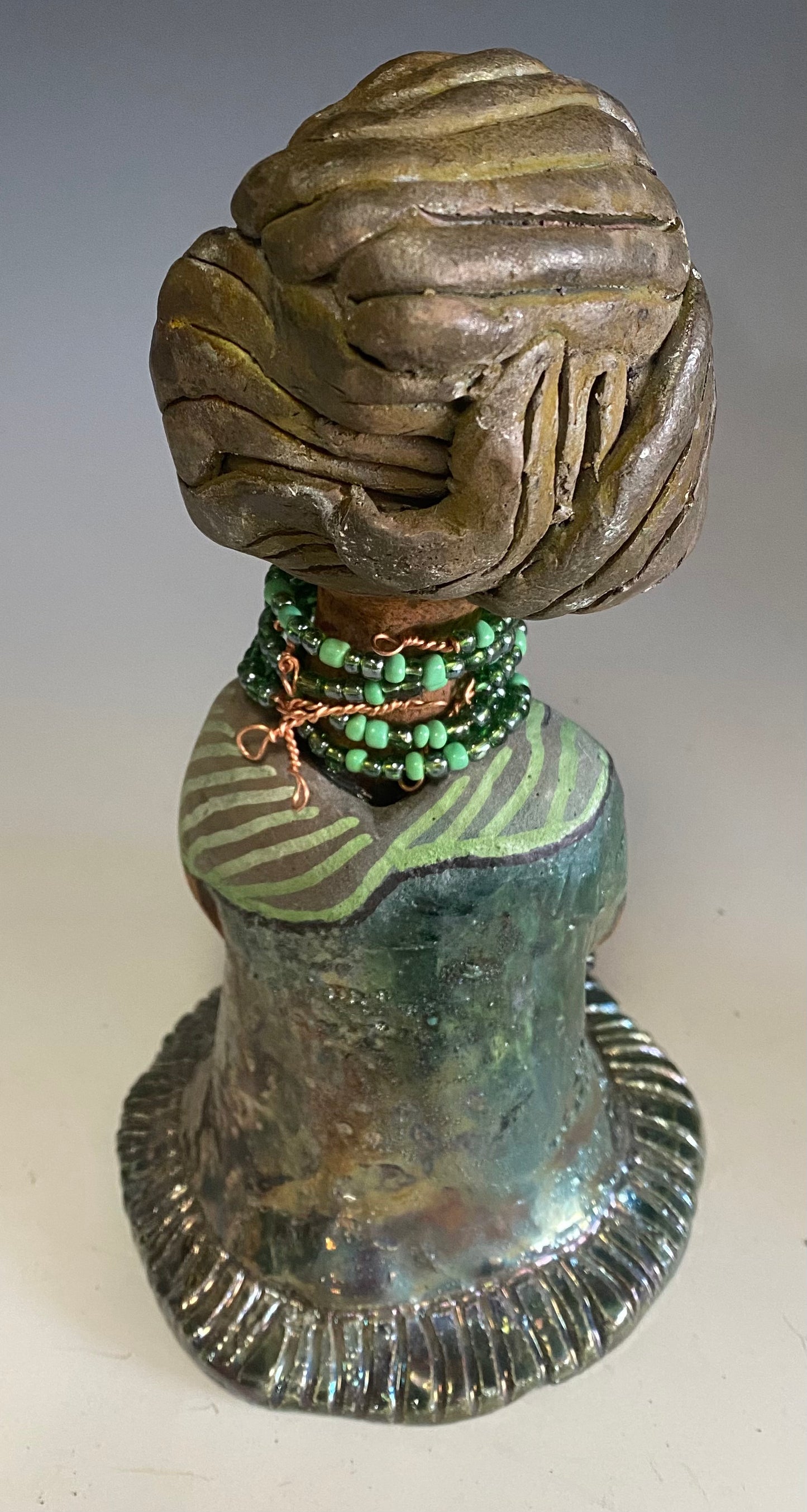 Meet Cora! Cora stands 9" x 5" x 6" and weighs 1.12 lbs. She has a lovely beige brown complexion with long etched braided clay hair. Cora’s long loving arms rest beside her copper green dress. Her dress is alligator green with a green coiled arm band. Cora wears a long string mixed green beaded necklace. Cora is a sophisticated lady that will grace your home.