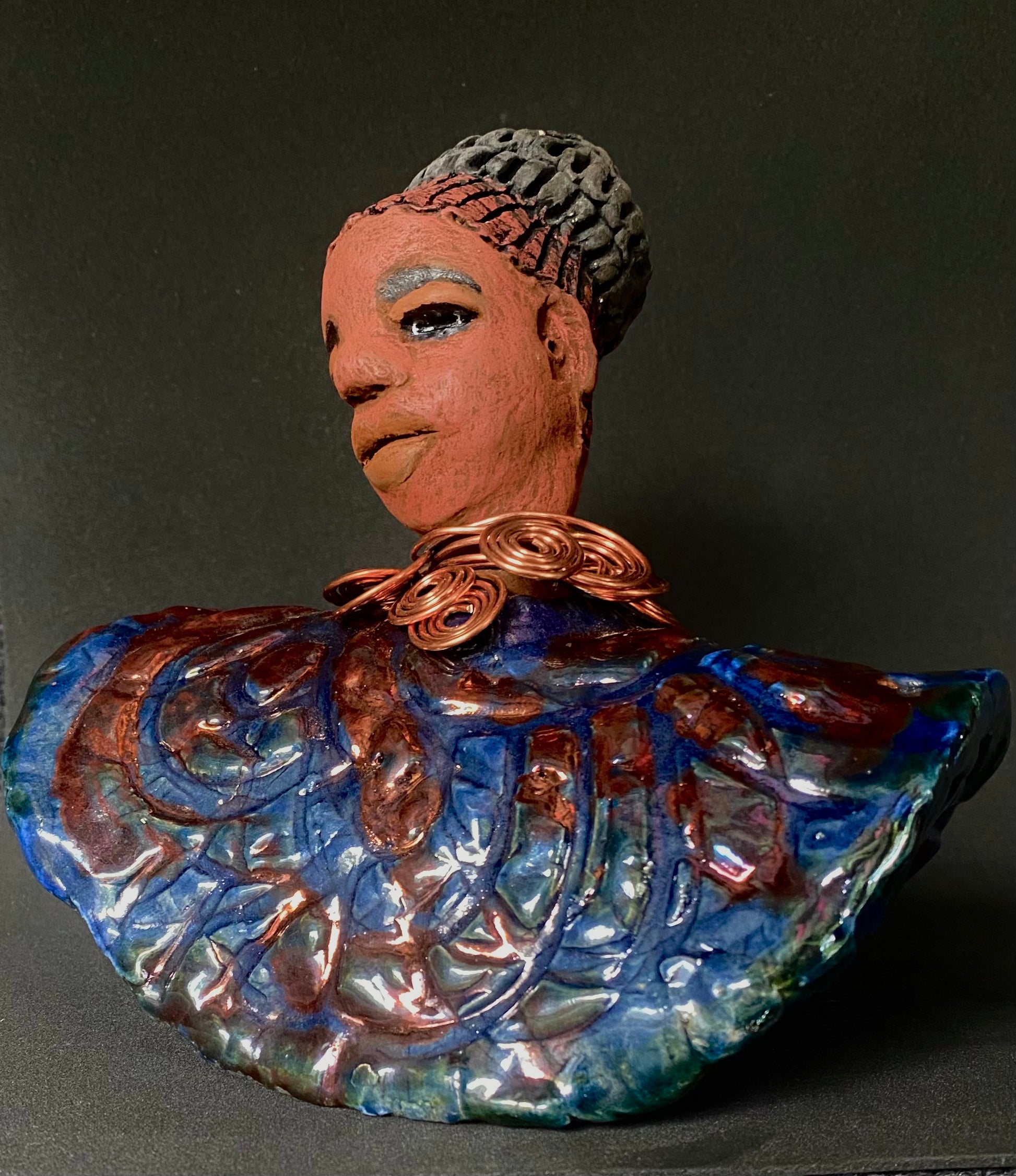 Meet Tia! Tia looks up with joy and anticipation! Tia stands 7" x 6" x 4" and weighs 1.90 lbs. She has a beautiful textured copper and blue robe. Tia has braided clay hair up in a bun. She has a honey brown complexion and soft light brown lips. Tia will make for great conversation in your home.  Free Shipping! Check out our Shipping and Return policy