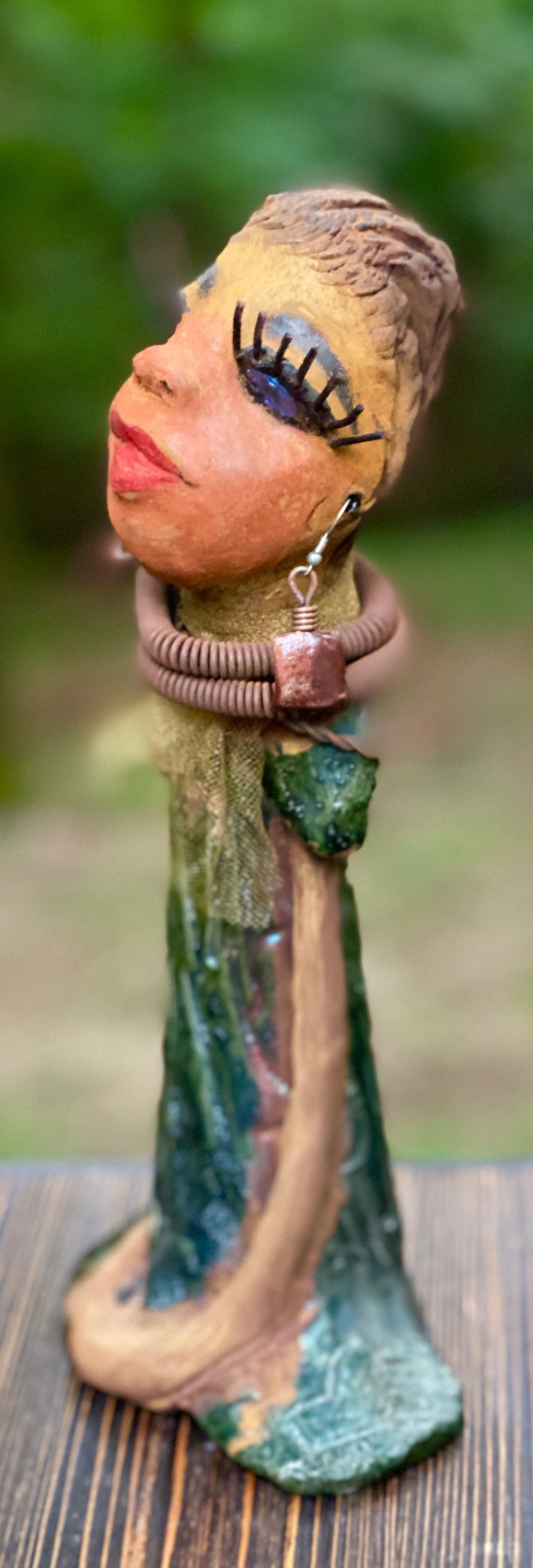 Meet Catera! Catera stands 10" x 4" x 4" and weighs 2.7 lbs. She has a lovely honey brown complexion with  reddish brown lips. She has a braided hairstyle.  Catera has a colorful metallic green antique copper glazed dress. She wears a yellow bead necklace with raku beads. Her long loving arms are resting at her side. With long lashes and eyes wide opened, Catera has hopes of finding a new home.