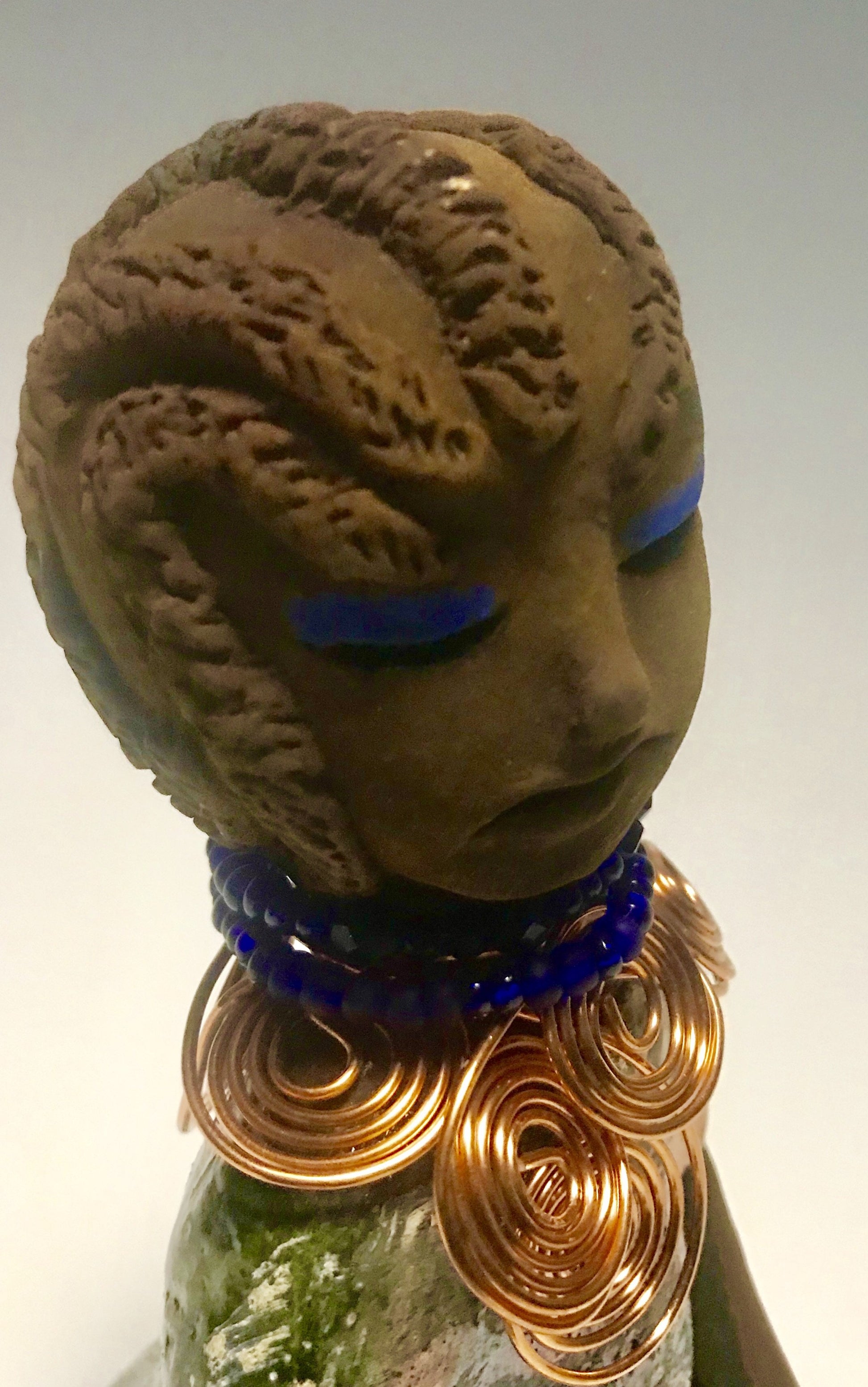 Anja stands 7" x 4" x 4" and weighs 1.01 lbs. She has a lovely honey brown complexion with  reddish brown lips. She has a braided hairstyle.  Anja has a colorful metallic green antique copper glazed dress. She wears a spiral copper wire necklace on top of a blue beaded collar. With eyes slightly opened, Anja has hopes of finding a new home.