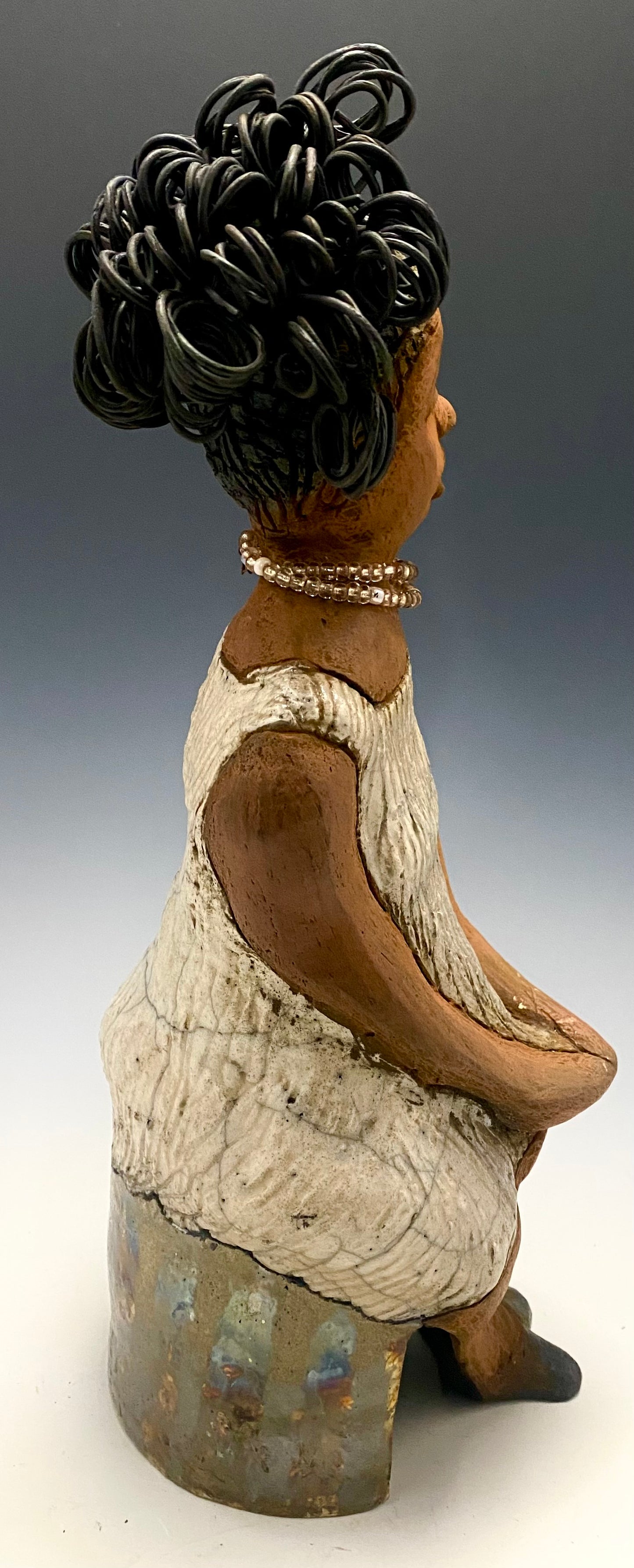 Meet Gwen! You know someone like Gwen. One who reaps elegance and sophistication! Gwen stands 13 " x 6" x 6" and weighs 3.07 lbs. She has a lovely brown complexion with over 5 feet of 16 gauge spiral coiled wire hair. Gwen's metallic off white dress is accented by a multicolored white and amber beaded necklace. Gwen’s straight face gives her a distinguished no nonsense look. Gwen sits with her hands rested as she awaits a special place to grace your home.