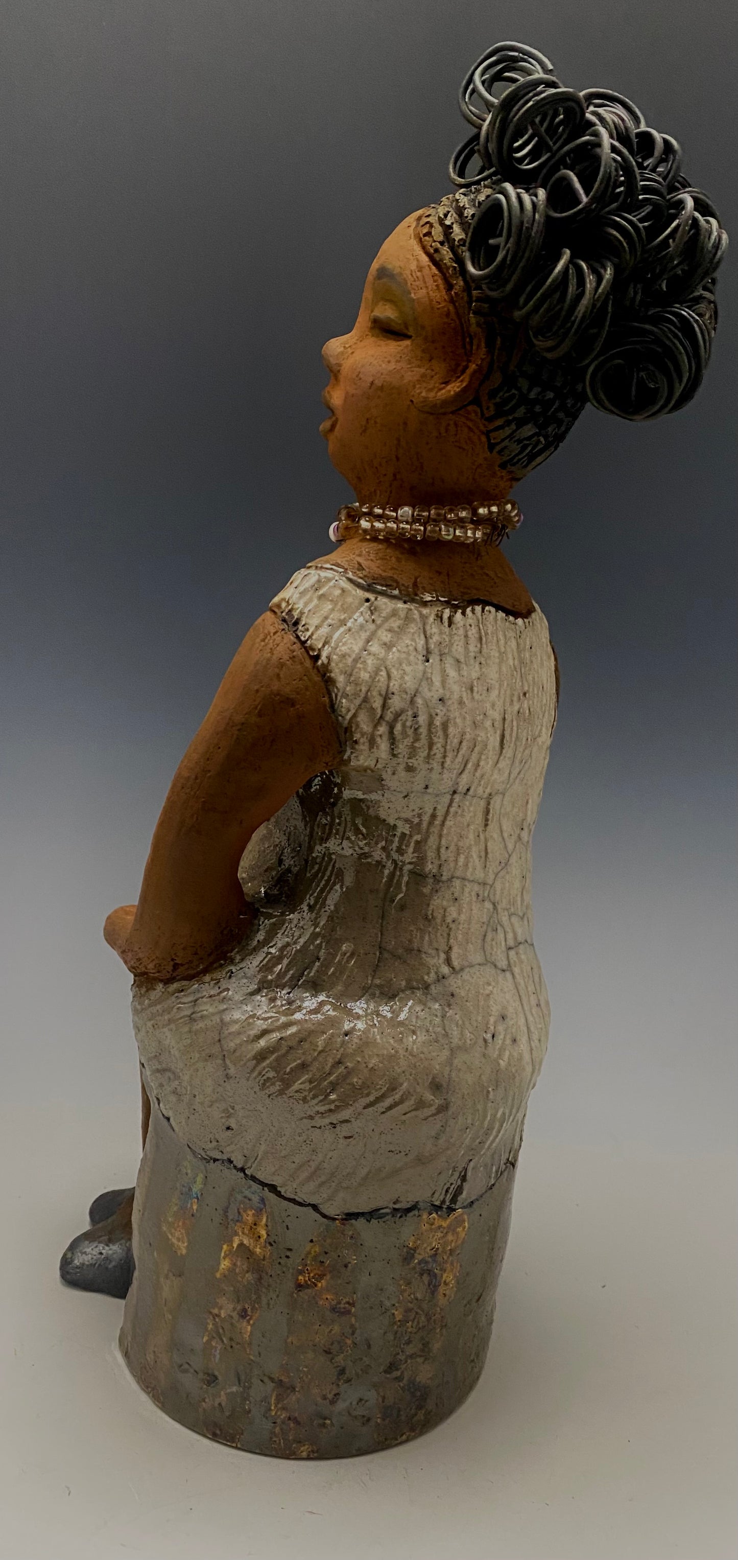 Meet Gwen! You know someone like Gwen. One who reaps elegance and sophistication! Gwen stands 13 " x 6" x 6" and weighs 3.07 lbs. She has a lovely brown complexion with over 5 feet of 16 gauge spiral coiled wire hair. Gwen's metallic off white dress is accented by a multicolored white and amber beaded necklace. Gwen’s straight face gives her a distinguished no nonsense look. Gwen sits with her hands rested as she awaits a special place to grace your home.