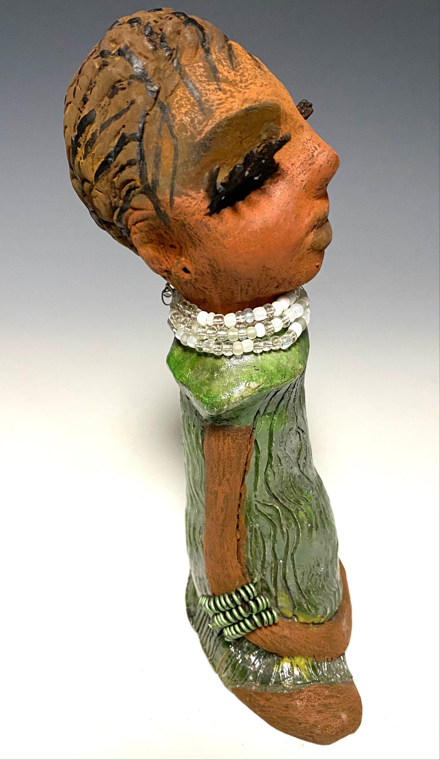 Meet Gracie! GracIe stands 9.5" x 6" x 3.5" and weighs 1.06 lbs. She has a lovely beige brown complexion with long etched braided clay hair. Gracie has a long white and clear beaded necklace. Gracie wears long stylish eyelashes Her long loving arms rest beside her copper green dress. Gracie is a sophisticated lady that will grace your home.