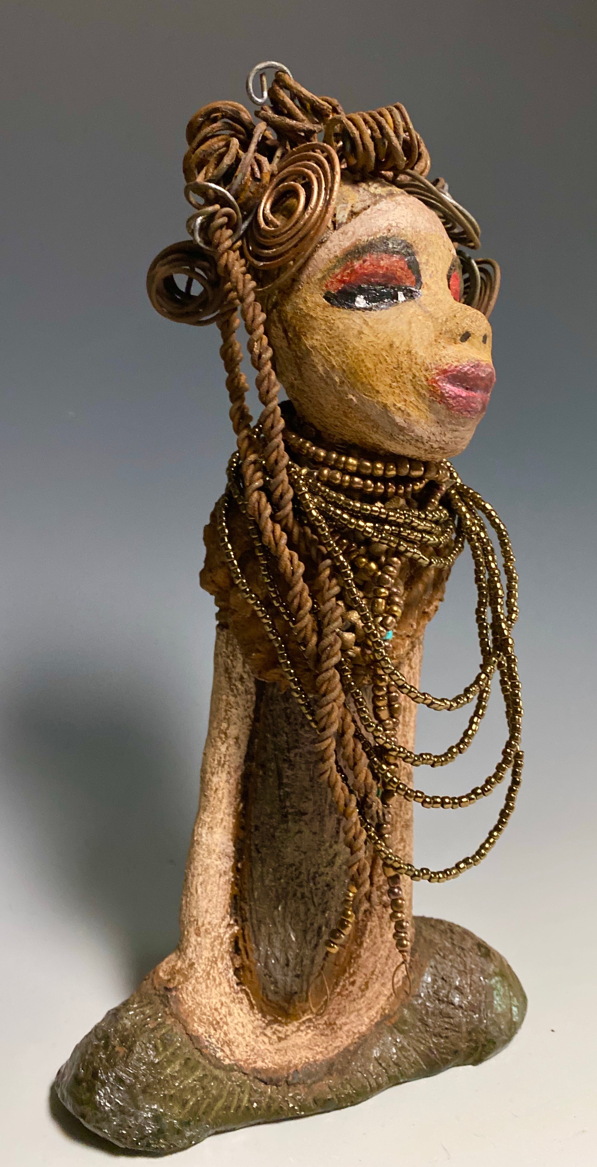 You will love having Cardi around! Cardi stands 11" x 5.5" x 3" and weighs 1.14 lbs. She has an awesome honey beige complexion. Cardi wears a dark metallic copper dress with a chocolate ruffle scarf.  She wears a multitude of amber colored hand strung beads. I do believe Cardi would like it if you gave her a space in your home!