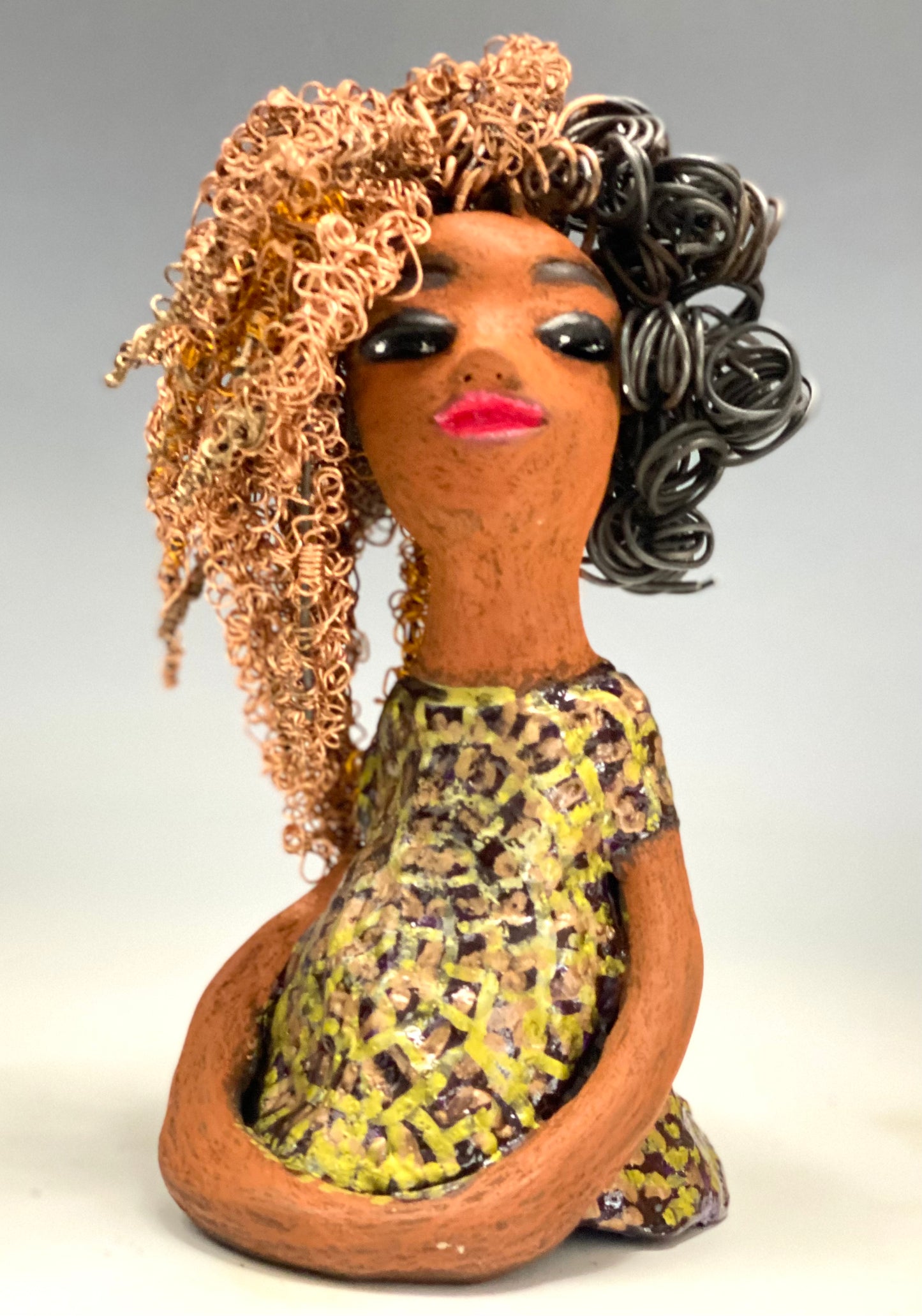 Meet Kalifa, an elegant figurine weighing 1.11 lbs and measuring 8" x 5" x 5.5". Her glossy spotted metallic copper glazed dress and long twisted wire locs are expertly crafted. With her honey brown complexion and red lips, she is sure to capture the attention of your guests. Her daring two-tone copper hair, which took 6 hours to create, adds to her beauty. Invite her into your home and spark meaningful conversations with her cheerful eyes and hopeful gaze.