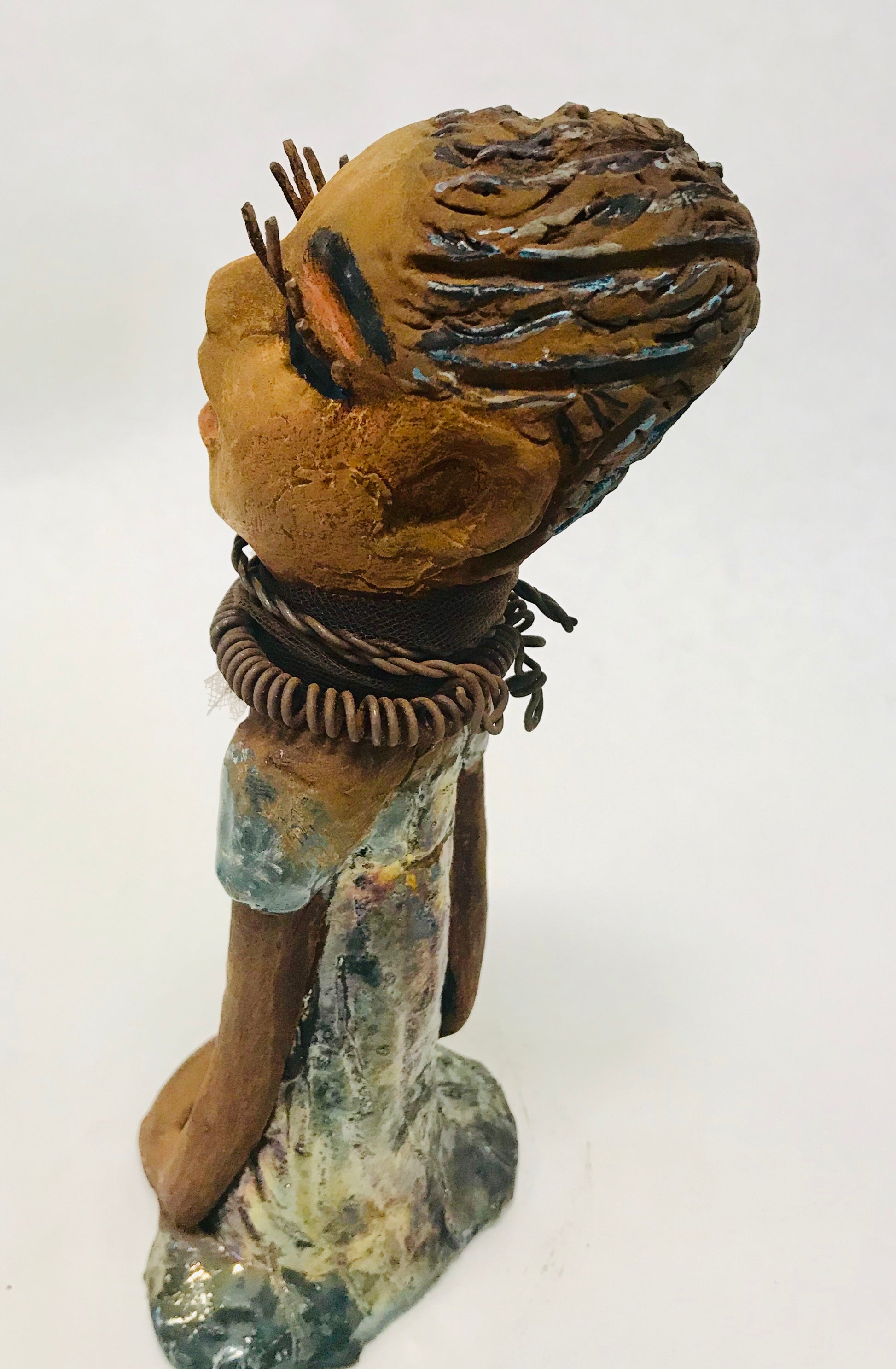 Sorbari stands 11" x 4" x 4" and weighs 2.5 lbs. She has a lovely honey brown complexion with  reddish brown lips. She has a braided hairstyle.  Sorbari has a colorful metallic green antique copper glazed dress. She wears two spiral wire necklaces. With long eyes slightly opened, Sorbari has hopes of finding a new home.