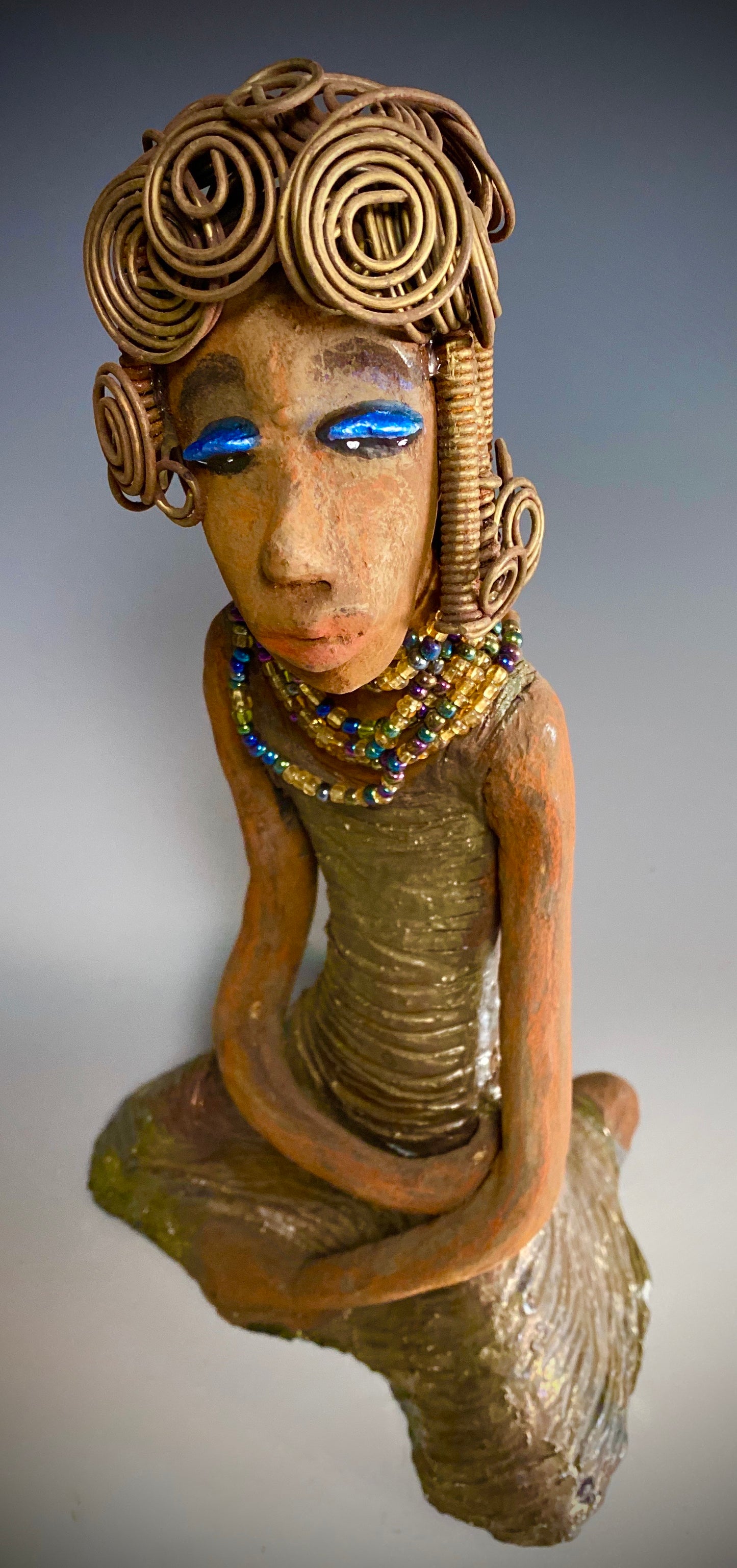 Meet Gloria! Gloria stands 12" x 7" x 5" and weighs 2.1 lbs. She has a lovely brown complexion. Her hair is gold and black. It took over 3 hours and 40 feet of coils and spirals of 16 gauge wire to complete! Gloria' coiled bangs hangs close to her slightly opened eyes. Wearing blue eye shadow, she has a multi colored dress with bright copper flashes to match. A string of iridescent amber colored beads surrounds her neck. With anticipation, Gloria wants to be with you.