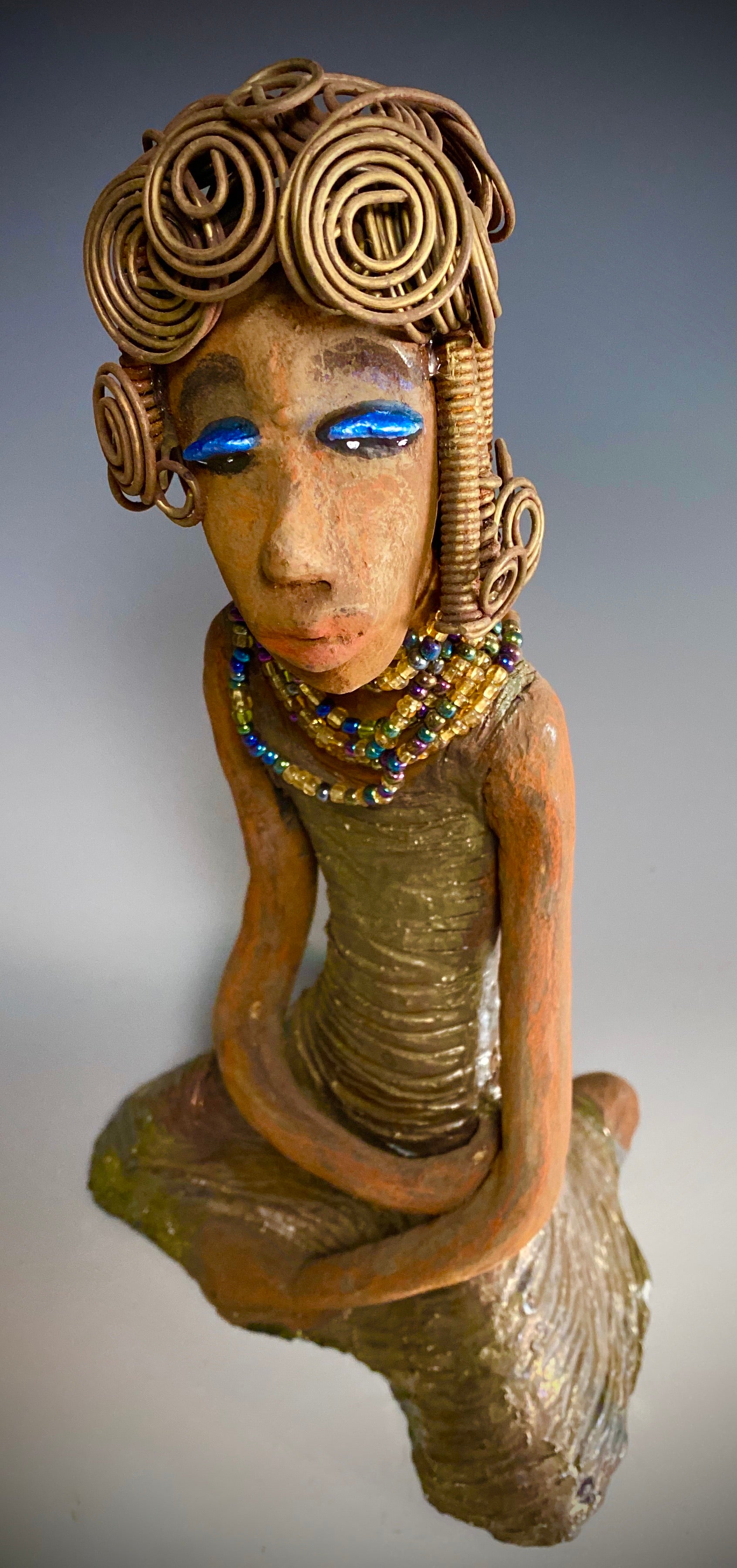 Meet Gloria! Gloria stands 12" x 7" x 5" and weighs 2.1 lbs. She has a lovely brown complexion. Her hair is gold and black. It took over 3 hours and 40 feet of coils and spirals of 16 gauge wire to complete! Gloria' coiled bangs hangs close to her slightly opened eyes. Wearing blue eye shadow, she has a multi colored dress with bright copper flashes to match. A string of iridescent amber colored beads surrounds her neck. With anticipation, Gloria wants to be with you.