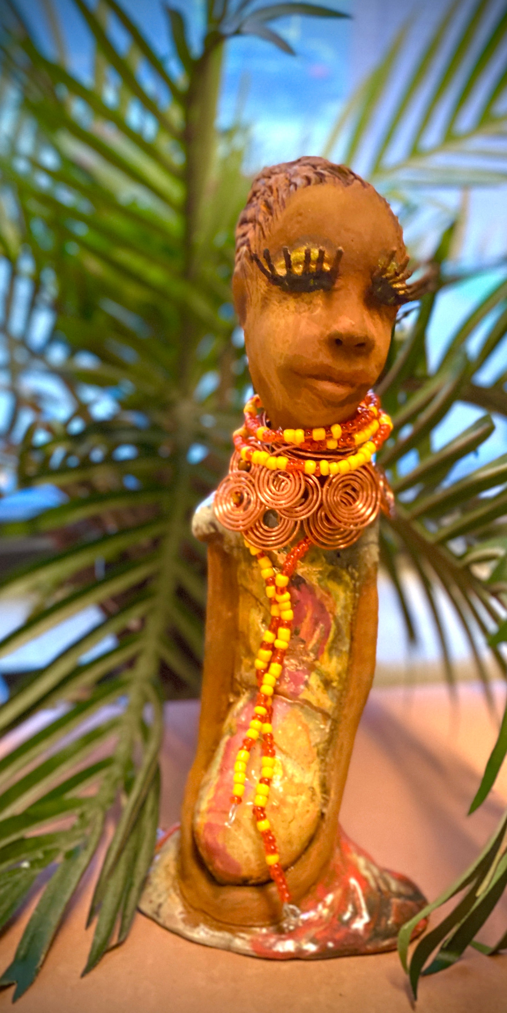 Reandra has a colorful reddish metallic green antique copper glazed dress. She wears a spiral copper wire necklace on top of a yellow red beaded collar. With eyes slightly opened, Reandra has hopes of finding a new home.