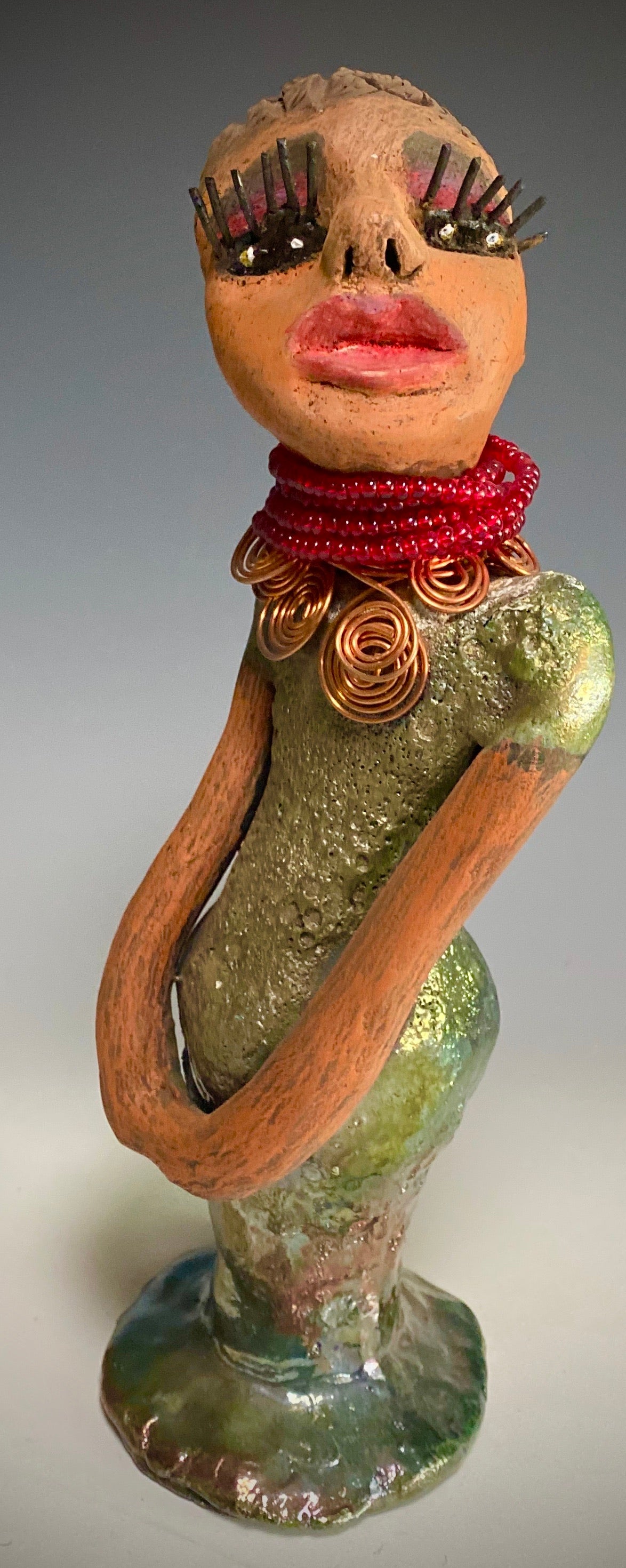 Meet Mona! Mona stands 11.5" x 4" x 4" and weighs 1.81 lbs. She has a lovely chocolate brown complexion with chocolate brown lips. She has a braided hairstyle. Mona has a colorful metallic antique copper glazed dress. She wears a spiral copper wire necklaces on top of a beaded ruby red collar. With long lashes and eyes wide opened, Mona has hopes of finding a new home. You may have noticed, Mona is the one of a few herdew sculptures that wears a face covering during this unprecedented time.