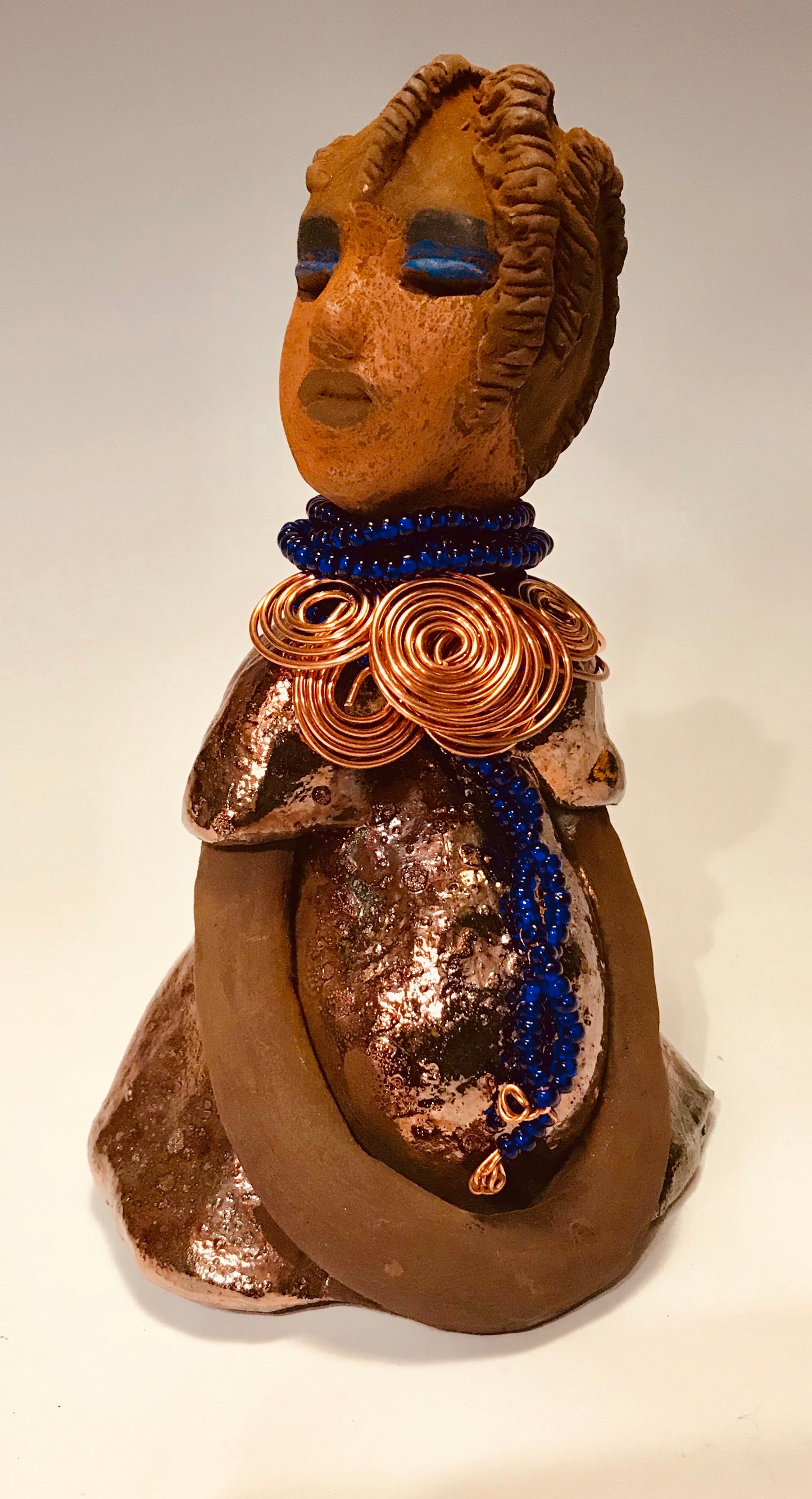 Ife stands 7.5" x 4" x 5" and weighs 1.02 lbs. She has a lovely honey brown complexion with reddish brown lips. She has a short braided hairstyle. Ife has a colorful metallic antique copper glazed dress. She wears spiral copper wire necklaces on top of an aqua blue beaded collar. Ife long loving arms rest at her side. With accent blue eye shadow and eyes wide opened, Ife has hopes of finding a new home.