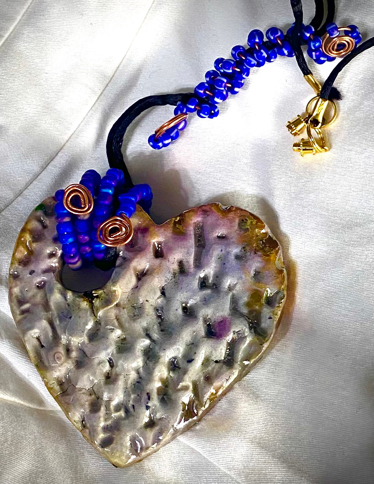 Have A Heart ! Each heart pendant is handmade with love! It is 3"x 3" and weighs approx. 3ozs. This pendant has a white metallic raku glazes that renders a unique translucent patina. The heart has a textured pattern . It holds a spiral of blue mini beads on a spiral copper wire. This pendant has a nice 12" black suede cord!