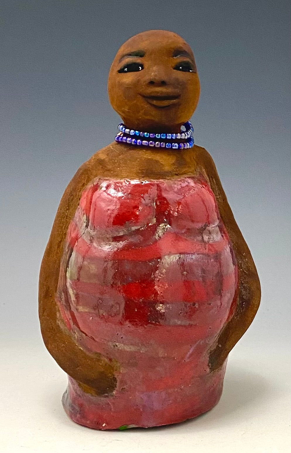 Meet Cora! Cora stands 9" x 7" x 4.5" and weighs  2lbs. She has a lovely honey brown complexion with lovely reddish brown lips. Cora wears a clay bald hairstyle. Cora has a striped heart red and gold glazed dress. She wears a blue beaded necklace. With eyes wide opened, Janae' has hopes of finding a new home.