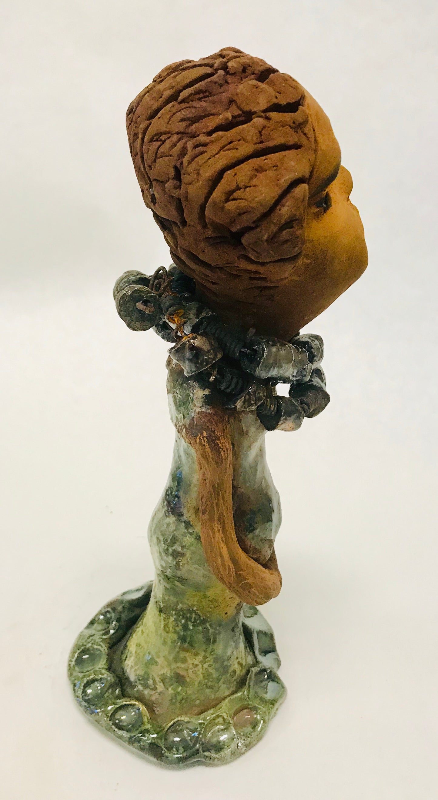 Greta stands 11" x 4" x 4" and weighs 2.5 lbs. She has a lovely honey brown complexion with reddish brown lips. She has a braided hairstyle. Greta has a colorful metallic green antique copper glazed dress. She wears two raku beaded necklaces. With long eyes slightly opened, Greta has hopes of finding a new home. Greta will attract and spark conversation with guest in your home!